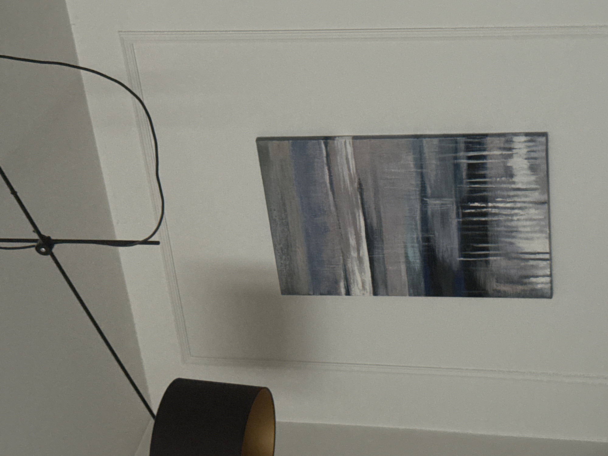 A framed abstract painting with horizontal and vertical brushstrokes in shades of blue, grey, and white hangs on a white wall within a framed paneled section. To the left of the painting, a portion of a black lampshade with a gold interior is visible. Above the painting and lampshade, a black electrical cord is suspended from the ceiling, attached by clips. The location is Unknown, Unknown.