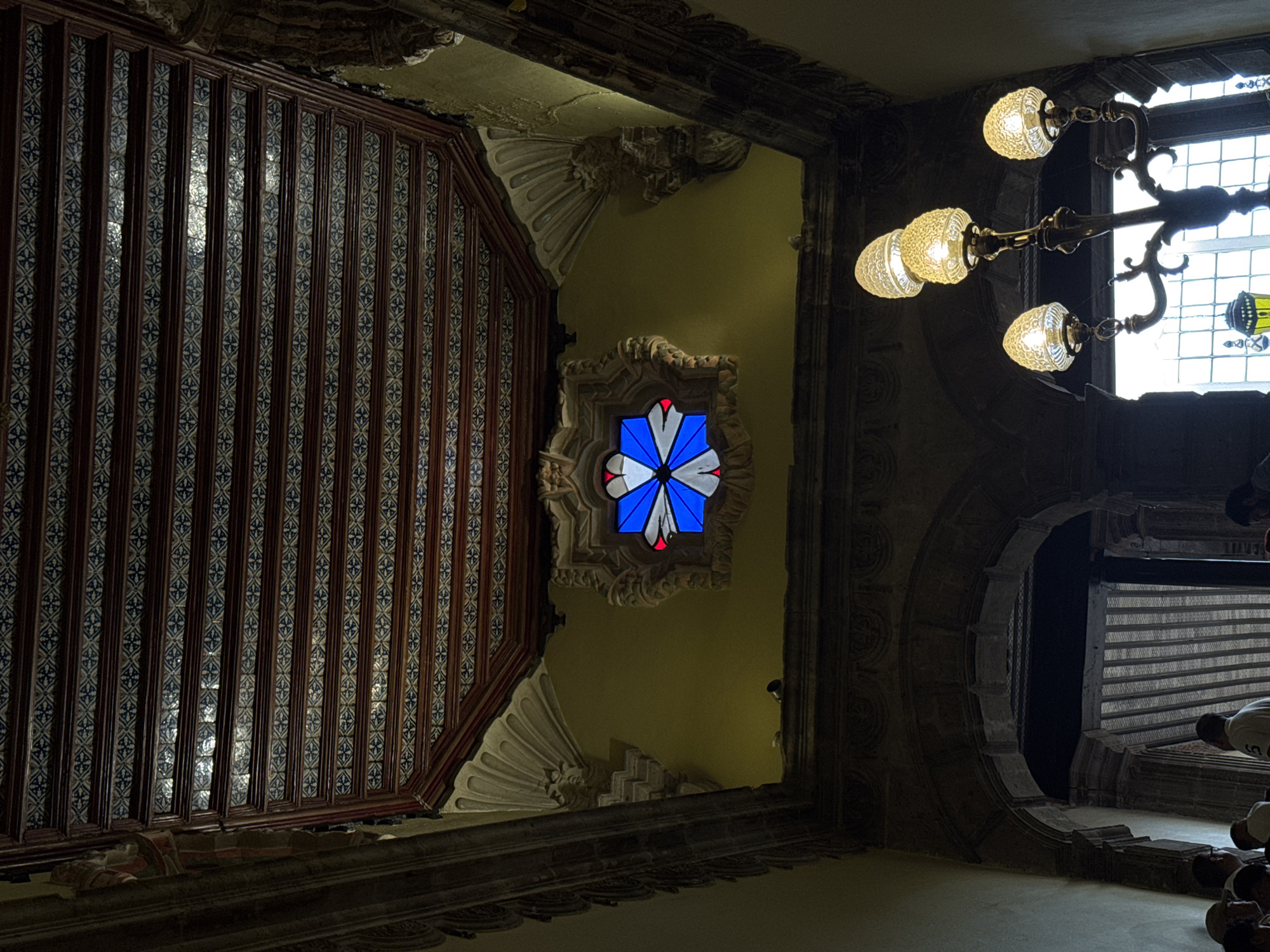 The image shows an interior view of a building in Mexico City, Mexico. The perspective is looking upwards towards the ceiling, featuring elaborate architectural details. On the left, there is a vertical structure with a pattern of brown frames and blue and white tiles. Adorning the ceiling are ornate carvings and a stained glass window with blue, white, and red colors. To the right, a vintage chandelier hangs from the ceiling, with several glass lamps illuminated. In the lower right corner, a circular archway provides a view of an adjacent area, where individuals are present.