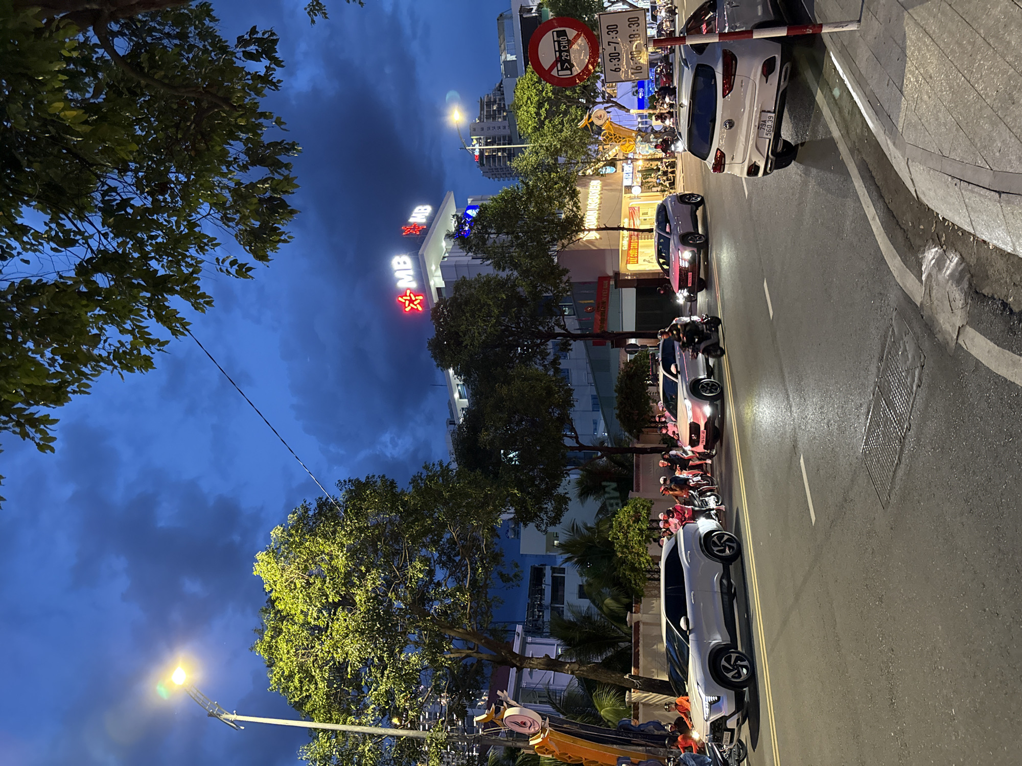 A street scene in Nha Trang, Vietnam, at dusk. The sky is a deep blue with scattered clouds, illuminated by streetlights and the neon glow of a building. Several cars and motorcycles are present on the road. In the foreground, a white SUV is parked on the right side of the road. A traffic sign with Vietnamese text and a circular symbol indicating a prohibition is visible on a pole. The background shows buildings, trees, and more vehicles. The overall atmosphere is that of a busy urban environment transitioning from day to night.