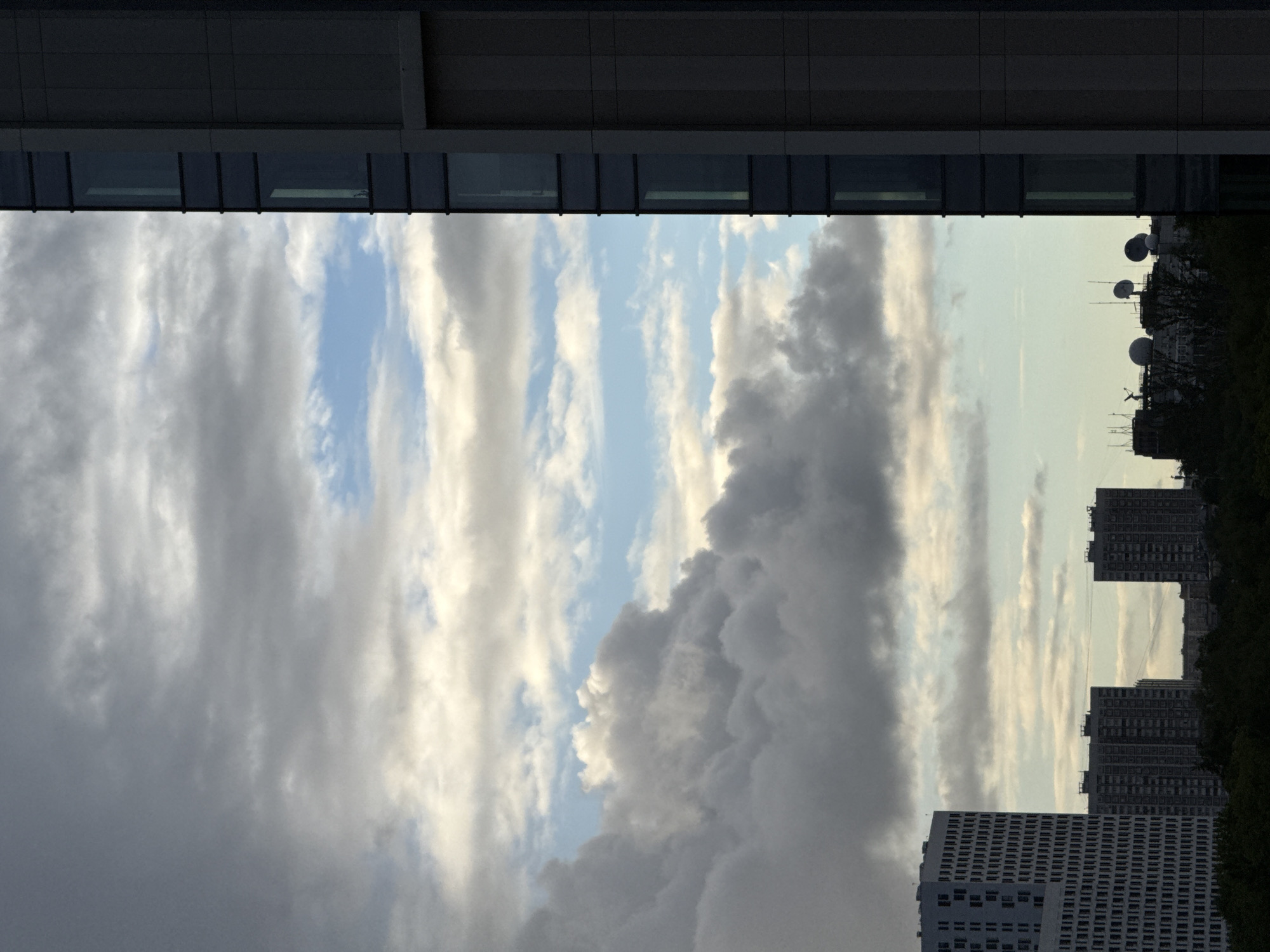 The image is taken from a low vantage point, looking up between two buildings. The top of the frame is occupied by a modern building facade with horizontal windows. Below this, the sky is visible, filled with a mix of dark and light clouds, some tinged with a golden color. In the bottom right of the frame, several high-rise buildings can be seen, characteristic of urban architecture. Based on the provided information, this scene is located in Khimki, Russia. The buildings and sky are the primary subjects in the image.