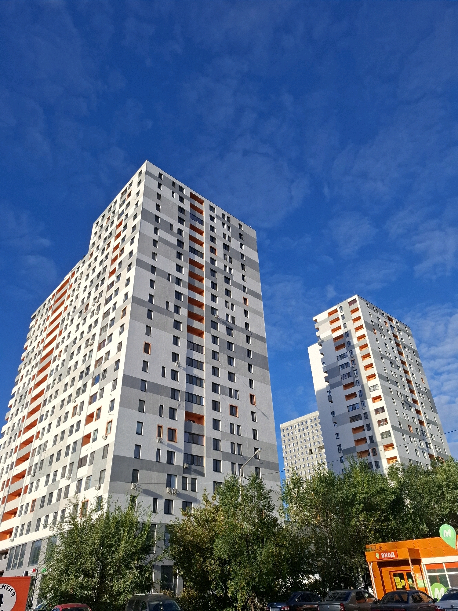 This is a low-angle, full-shot photograph of a modern residential complex under a bright blue sky with scattered clouds. The buildings are tall, multi-story apartment blocks with a contemporary design, featuring a facade of white, gray, and bright orange elements. The orange accents are prominent, particularly on the balconies and window frames, adding a vibrant contrast to the otherwise neutral building colors. Numerous windows are visible on each building. In the foreground, there are green trees and a row of parked cars. To the right, a bright orange storefront with "ВХОД" (Entrance) written in Russian above the door is visible, suggesting a commercial establishment at the base of one of the buildings. The overall impression is an urban residential area. This scene is consistent with a view of the "European Premier" residential complex in Tyumen, Russia, known for its modern architecture and colorful facade elements.
