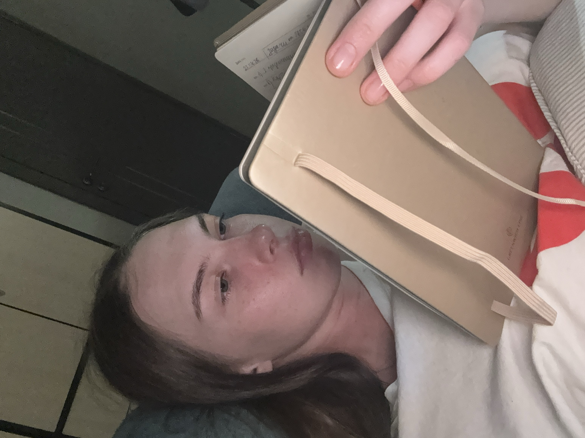 A young woman with light brown hair is looking down at a notebook. She is lying in bed, propped up by pillows, and is wearing a white t-shirt. The room appears to be dimly lit, with a closet door visible in the background. The notebook has a light brown cover with a beige elastic band and ribbon bookmark. The woman's face is partially obscured by the notebook. There are no identifiable features that would suggest a specific location within the city of Perm, Russia.