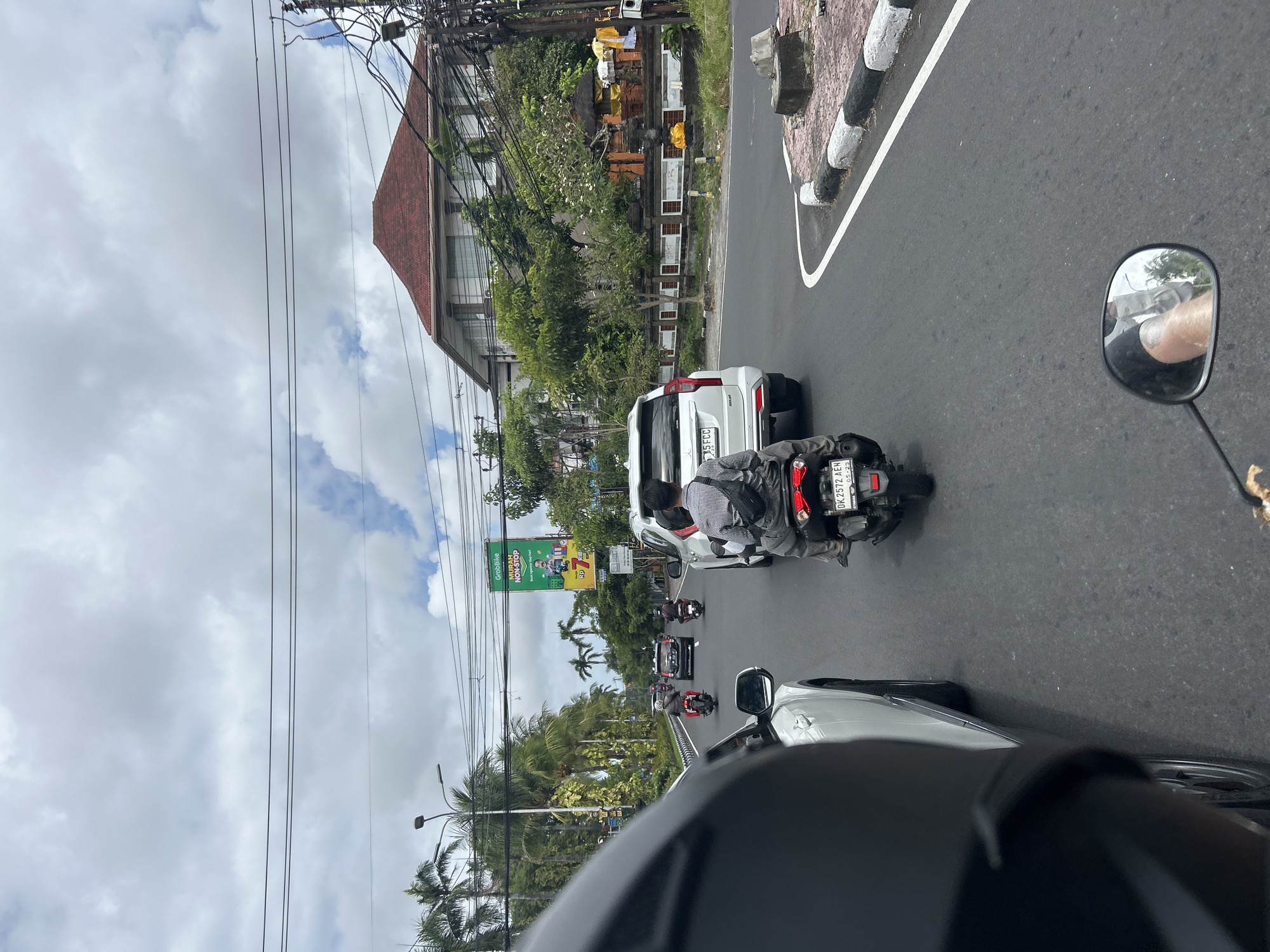 The image depicts a street scene in Denpasar, Indonesia, likely on a cloudy day. In the foreground, the left side of a black helmet and the side of a white car are visible, suggesting the photo is taken from a vehicle. Further down the road, several motorcycles and a white SUV are in traffic. On one motorcycle, a person wearing a plaid shirt and a black fanny pack is riding. The white SUV has a license plate that reads "DK 1572 AEN". The background shows a street lined with palm trees and other vegetation. A building with a red tiled roof and several stories is visible, as is a billboard for "GrabBike". Electrical wires crisscross above the street. To the right, a small temple structure with orange walls and white accents is present, adorned with yellow fabric. The road has white and black painted curbs.