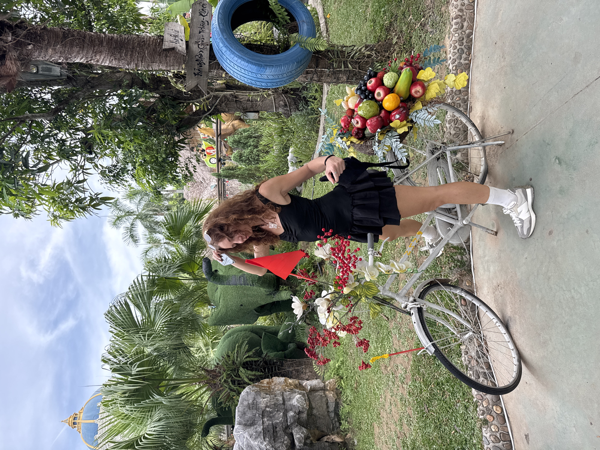 A woman with curly brown hair, wearing a black dress and white sneakers, stands with one foot on the pedal of a white bicycle. The bicycle's front basket is overflowing with a colorful arrangement of fruits and flowers, including apples, oranges, grapes, and red berries. A red flag is affixed to the bicycle's handlebars. In the background, green, topiary-like elephant sculptures are visible, along with palm trees and a large building with a blue dome. A blue tire is mounted on a tree to the right. The scene appears to be an outdoor park or garden.
