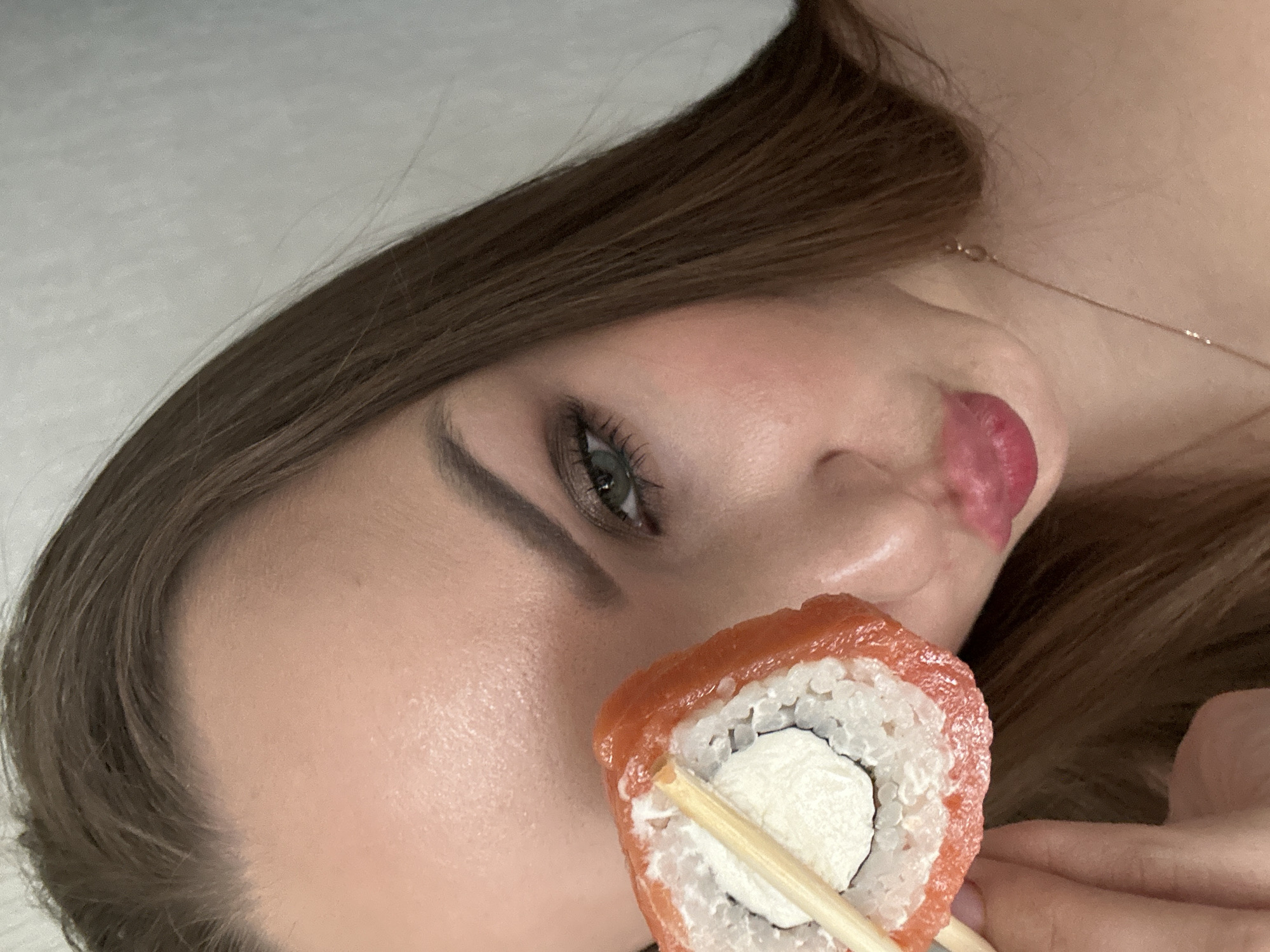 A close-up, canted image shows a young woman holding a piece of sushi with chopsticks. The sushi appears to be a California roll, wrapped in salmon. The woman has light skin, green eyes, brown hair, and a thin gold necklace. Her lips are parted slightly. The background is a plain, light color. The location of the image in the city of Chaikovsky, Russia is not evident from the image.