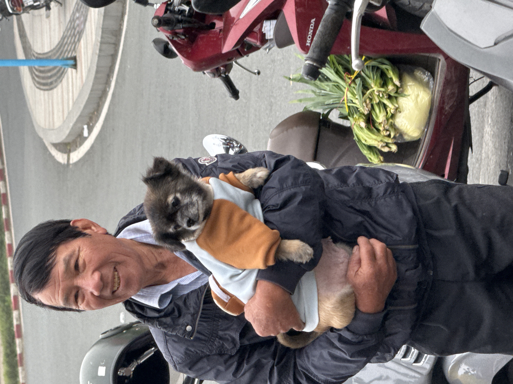 An elderly East Asian man with black hair and a light blue collared shirt under a dark blue jacket stands outdoors, holding a small, fluffy dog with dark fur. The dog is wearing a light blue and orange sweater. The man is smiling and looking slightly to his left. He is standing next to a dark helmet on a motorcycle. In the background, a red motorcycle is visible, with a bunch of green vegetables wrapped in plastic in its compartment. The scene appears to be on a paved street with a sidewalk and a curb. The lighting suggests it is daytime.