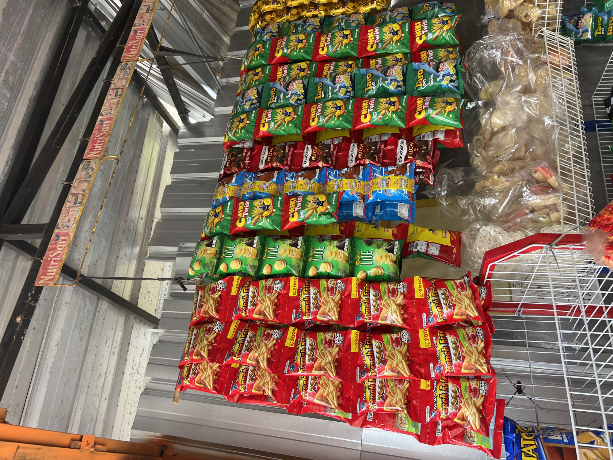 The image shows a roadside stall in Rendang, Indonesia, selling a variety of snacks and drinks. The stall is covered by a corrugated metal roof, supported by metal beams. Hanging from the roof are numerous rows of brightly colored snack bags, predominantly featuring chips and crackers. Prominently displayed are bags of "French Fries" potato chips and "Chiki Twist" snacks in various flavors like seaweed. Other visible snack brands include "Taro net" and "Chitato Lite". Below the hanging snacks, a white metal shelf holds larger bags of what appear to be crackers and snacks. A sign advertising "NutriSari" is visible hanging from the roof structure. The overall scene depicts a bustling local market stall typical of a small Indonesian town.
