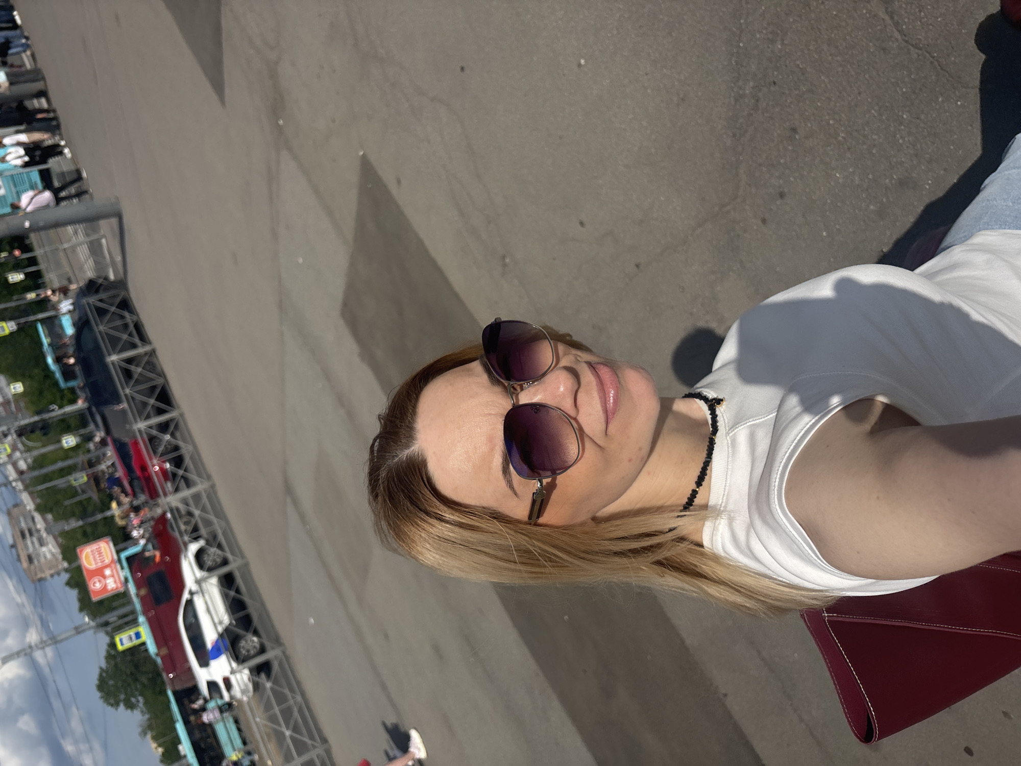 The image features a fair-skinned woman taking a selfie outdoors under bright sunlight, angled to show her upper body and the surrounding environment. She has long, straight blonde hair and wears dark, aviator-style sunglasses that obscure her eyes. Her lips are slightly parted in a neutral expression, adorned with a light pink gloss. She is wearing a plain white short-sleeved t-shirt and a black beaded necklace. A deep red handbag is visible under her left arm.
The setting is an urban outdoor space, likely a street or a parking area adjacent to a road with multiple lanes of traffic. The sun is high, casting distinct, elongated shadows on the light-colored pavement, indicating it is likely mid-day or afternoon on a clear, sunny day.
In the background, numerous vehicles are visible, including several cars parked or in motion. Notable among them are a red car and a white car with blue stripes, resembling an emergency or service vehicle. The background also shows parts of buildings and some trees.
Prominently visible textual cues include a "Burger King" restaurant sign. Below it, a red sign with Cyrillic text displays "ТЦ" (likely an abbreviation for "Shopping Center" or "Shopping and Entertainment Complex") and "360 m," indicating a distance. A green street sign with a white arrow is also present. These details, particularly the Cyrillic text, confirm the location context of Saint Petersburg, Russia. The overall scene depicts a bustling urban environment.