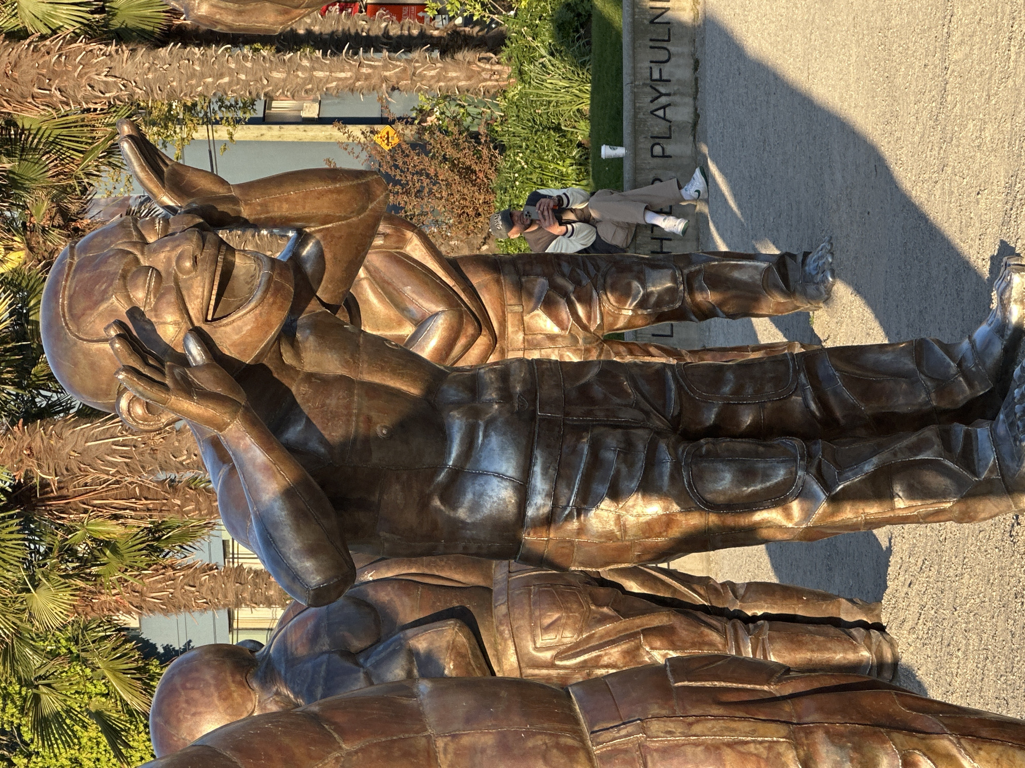 The image captures a large bronze sculpture in an outdoor public space, likely a park or sculpture garden in Vancouver, Canada. The sculpture consists of several stylized human figures, cast in a textured bronze, depicting joyful or playful interaction. One figure in the foreground appears to be laughing heartily, with hands raised to its head. The figures are dressed in what resembles contemporary clothing, including jackets and trousers with visible seams and pockets.
In the background, a person is seated on a low concrete wall, engaged with their phone. They wear a cap, a light-colored jacket, and light trousers with white sneakers. A white cup rests on the wall beside them. The scene is bathed in the warm, golden light of the late afternoon, casting long shadows across the paved ground, indicating clear, sunny weather.
Palm trees and other green foliage frame the background, suggesting a mild climate. A significant detail is the word "PLAYFULNESS" inscribed on the concrete wall behind the seated individual, reinforcing the thematic element conveyed by the sculpture's expressive figures. The overall impression is one of art integrated into a vibrant public environment.