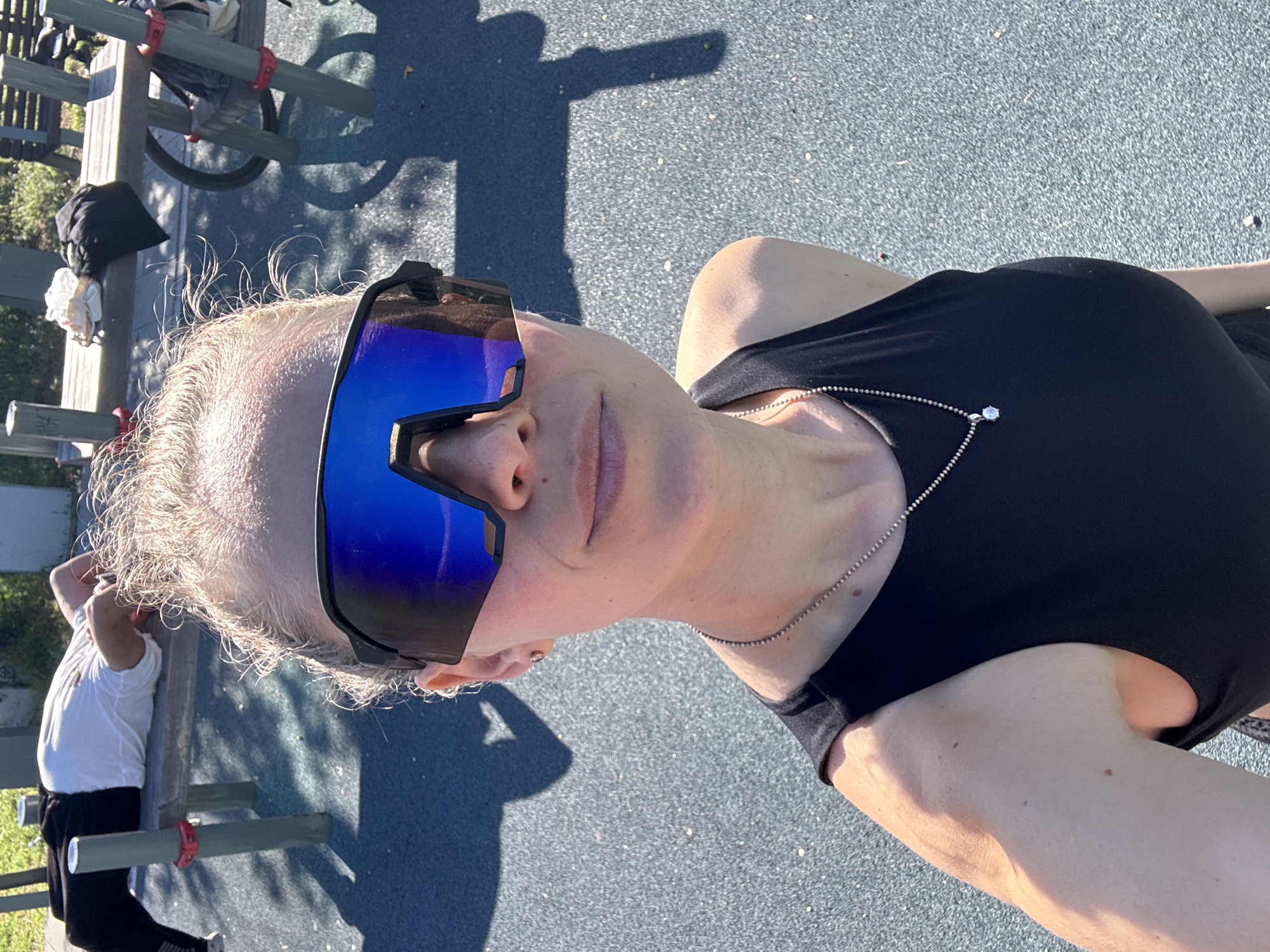 A woman wearing large mirrored sunglasses and a black tank top is posing in a selfie. She is wearing a silver necklace. The background shows a park with benches and a bicycle. The location is likely a park in a city given the urban infrastructure. It is not possible to confirm the specific location of городское поселение Щелкино, Ukraine from this image.