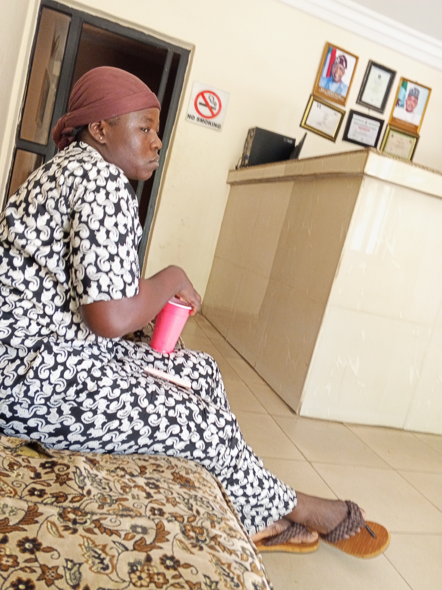 A woman with a brown headscarf and a black and white patterned outfit is seated on an ornate cushion. She's holding a red plastic cup. A "No Smoking" sign is on the wall behind her. Several framed portraits and certificates hang above a reception desk. The location is in the city of Unknown, Unknown.