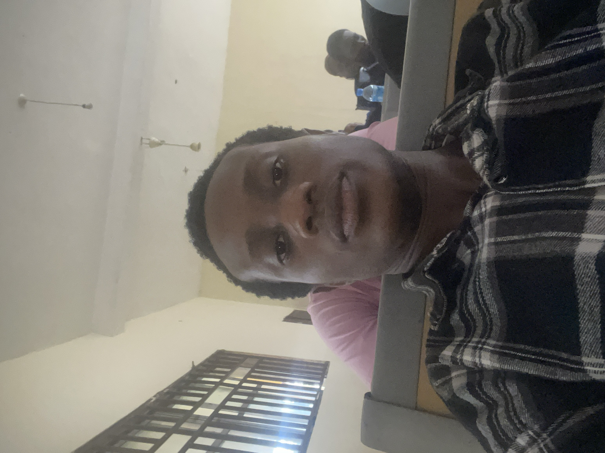 A young Black male is taking a selfie in what appears to be a classroom or lecture hall. He is wearing a black and white plaid shirt. In the background, there are other students seated, and a blue water bottle is visible on a desk. A window with a barred grate is on the left side of the image, and bare light bulbs hang from the ceiling. The overall lighting suggests it is daytime. The location is identified as Ihiagwa, Nigeria.