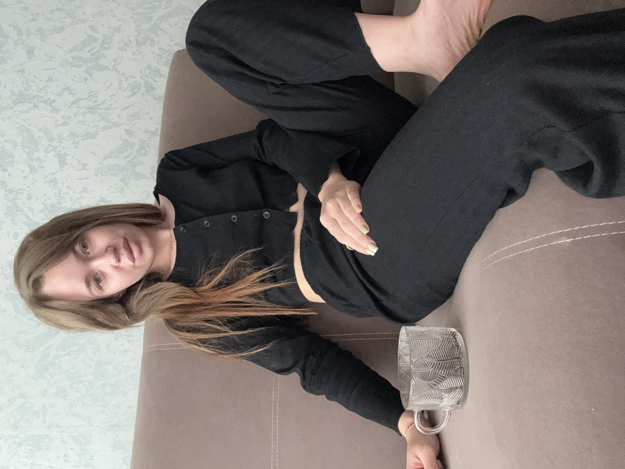 A woman with light brown hair is sitting on a brown sofa, looking at the camera. She is wearing a black long-sleeved crop top and black pants. To her left is a clear glass mug with a patterned design. The background is a wall with a light blue and white textured wallpaper. The image appears to be taken indoors, likely in a residential setting. There is no specific information available about the location in the city of Chaikovsky, Russia.