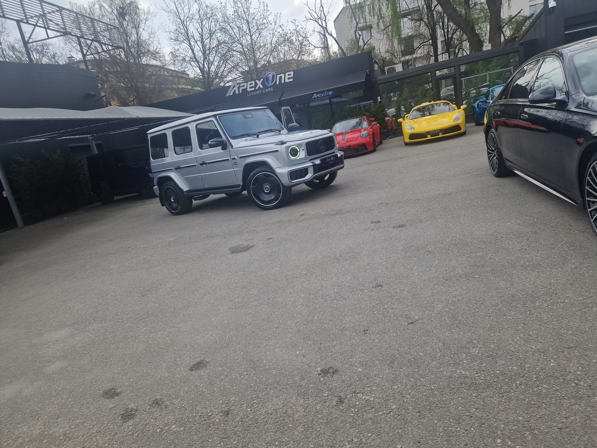 This image depicts an outdoor scene at a luxury car dealership called "APEX One LUXURY CARS" on an overcast day. The foreground and midground are dominated by a paved, grey asphalt parking lot.
The central subject is a silver Mercedes-Benz G-Class (G-Wagen) SUV, positioned in the mid-left. It features prominent round LED headlights, large black multi-spoke wheels, and a "V8 BITURBO" badge on its side. The text "APEX One" is visible on its front bumper. Behind it, partially obscured by a dark awning, another dark-colored SUV or large vehicle is parked.
To the right of the G-Wagen, a striking red sports car, possibly a Porsche 911 GT3 RS, is parked, angled slightly towards the left. Further to the right and slightly behind the red car, a bright yellow sports car, resembling a Ferrari 458, is also parked facing left. A black luxury sedan, likely a Mercedes-Benz, is partially visible on the far right, with its front section cropped out of the frame. Through the glass windows of the dealership building, a blue car can be faintly seen inside.
The dealership building itself has a modern, dark facade with expansive glass display windows. A prominent white sign above the entrance reads "APEX One LUXURY CARS." A smaller "APEX One LUXURY CARS" sign is also visible within one of the display windows.
The background features several trees, some with bare branches and others with early spring foliage, suggesting the season. A multi-story building is visible behind the trees on the right. The sky is uniformly grey and overcast, indicating diffused lighting. No people are present in the image.