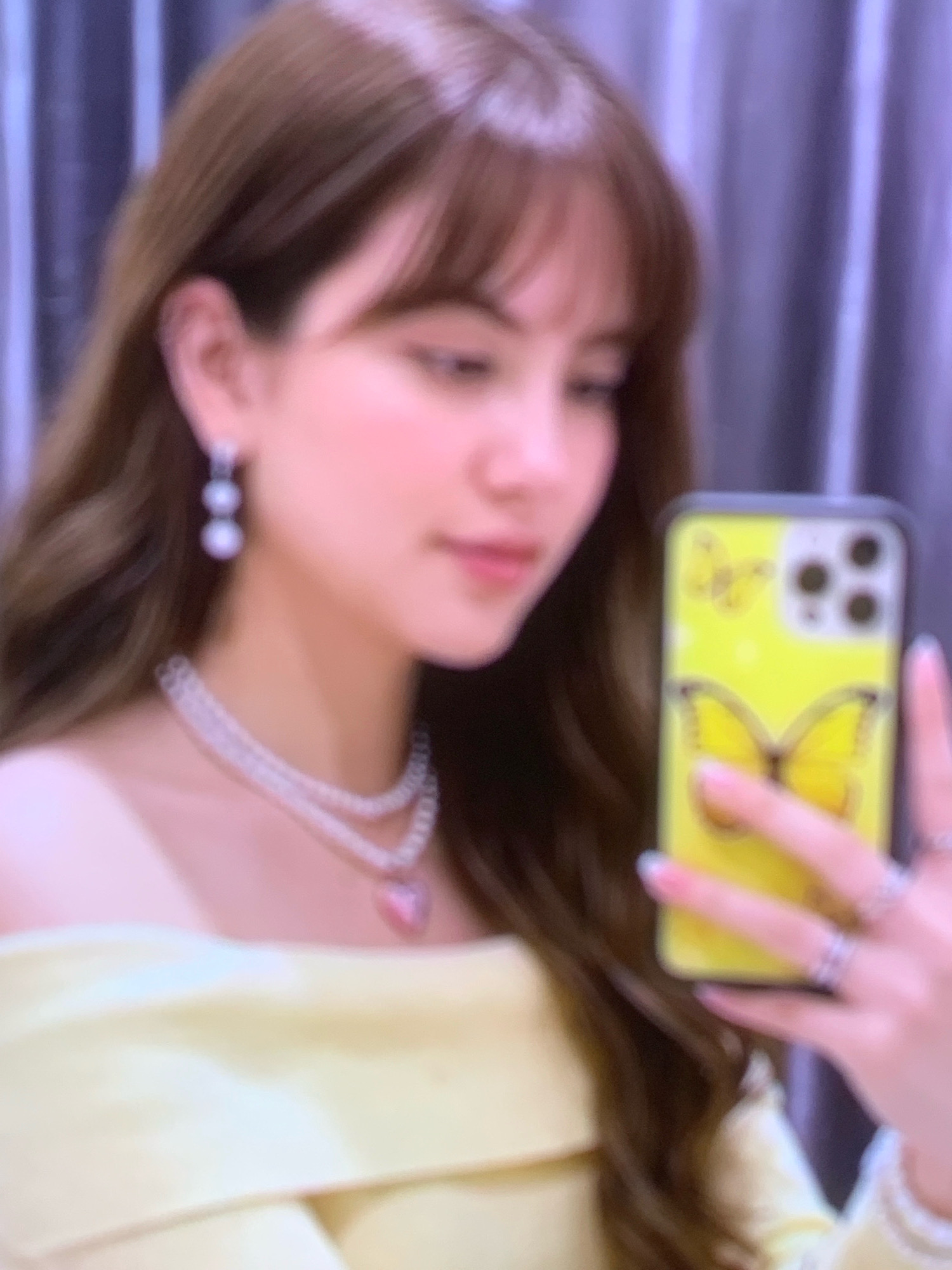 A person with long, brown hair and bangs is holding a yellow smartphone with a butterfly design in their right hand, taking a selfie. They are wearing pearl earrings, a multi-layered pearl necklace with a pink heart pendant, and a light yellow off-the-shoulder top. The background appears to be a curtain. The image is blurry and has a pinkish hue.