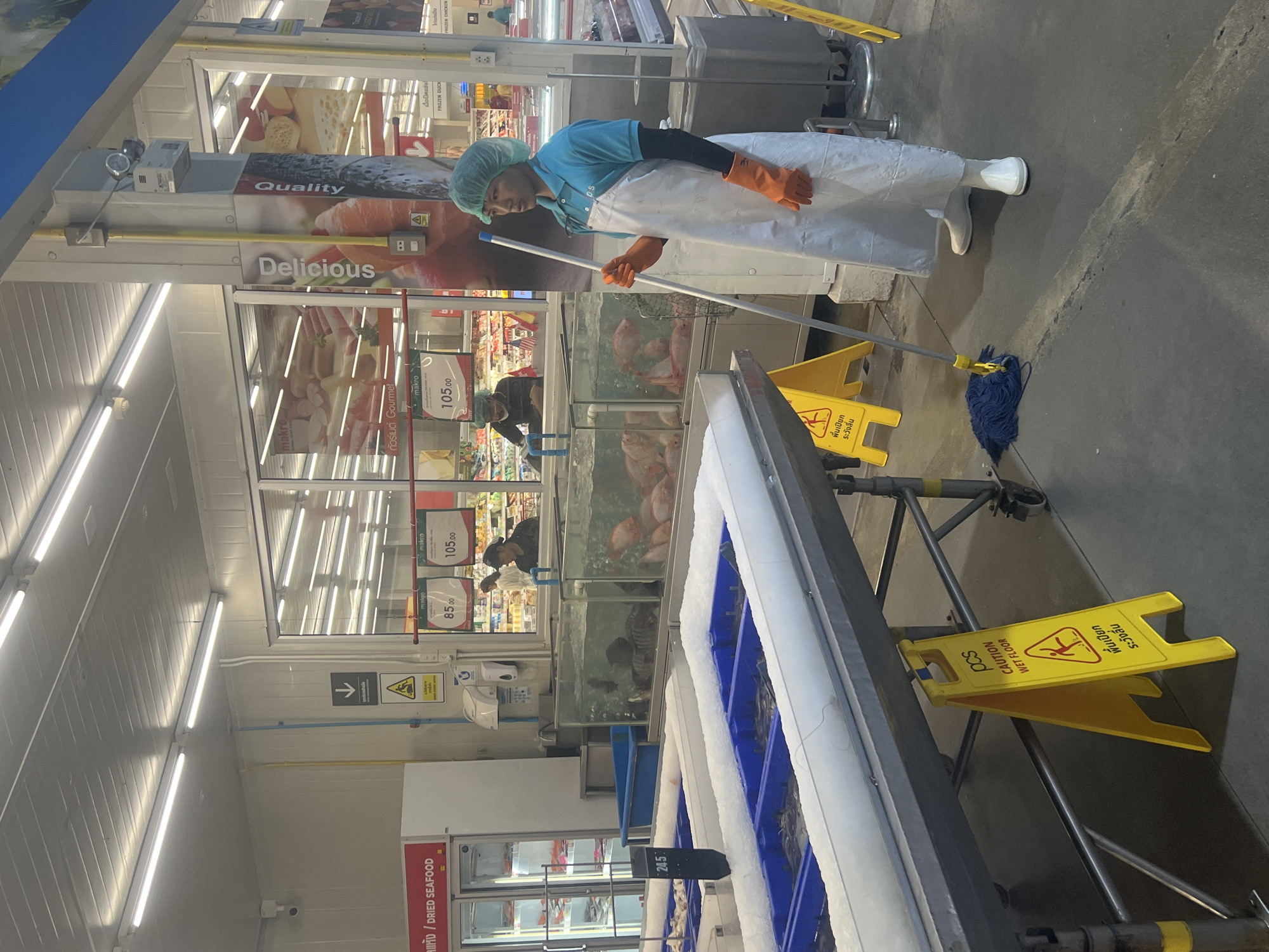 A male worker in a blue shirt, white apron, hairnet, and orange gloves is mopping the floor in a grocery store in Ko Samui District, Thailand. The floor is dark grey, and a wet floor sign is visible. The worker is holding a mop with a blue head. In the background, there are large refrigerated display cases filled with seafood on ice. Further back, there are large tanks containing live fish, and shelves stocked with groceries. The store appears to be well-lit with fluorescent lighting.