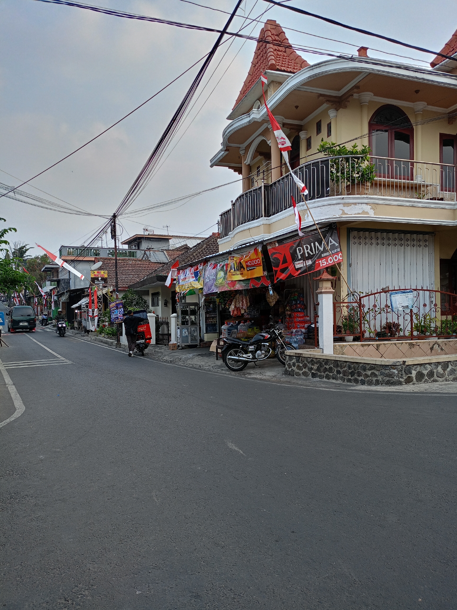 A street scene in Batu, Indonesia features a two-story building with yellow walls and a red tiled roof, adorned with Indonesian flags. A balcony with a black metal railing extends from the second story. Several shops line the street, with awnings and banners displaying various advertisements. Two men are seen near a red tricycle. A motorcycle is parked on the sidewalk, and another motorcycle is in motion on the street. A dark green van is also present on the road. Overhead, a tangle of power lines crisscrosses the sky. The street itself is paved, with white lines marking a lane. The weather appears overcast.