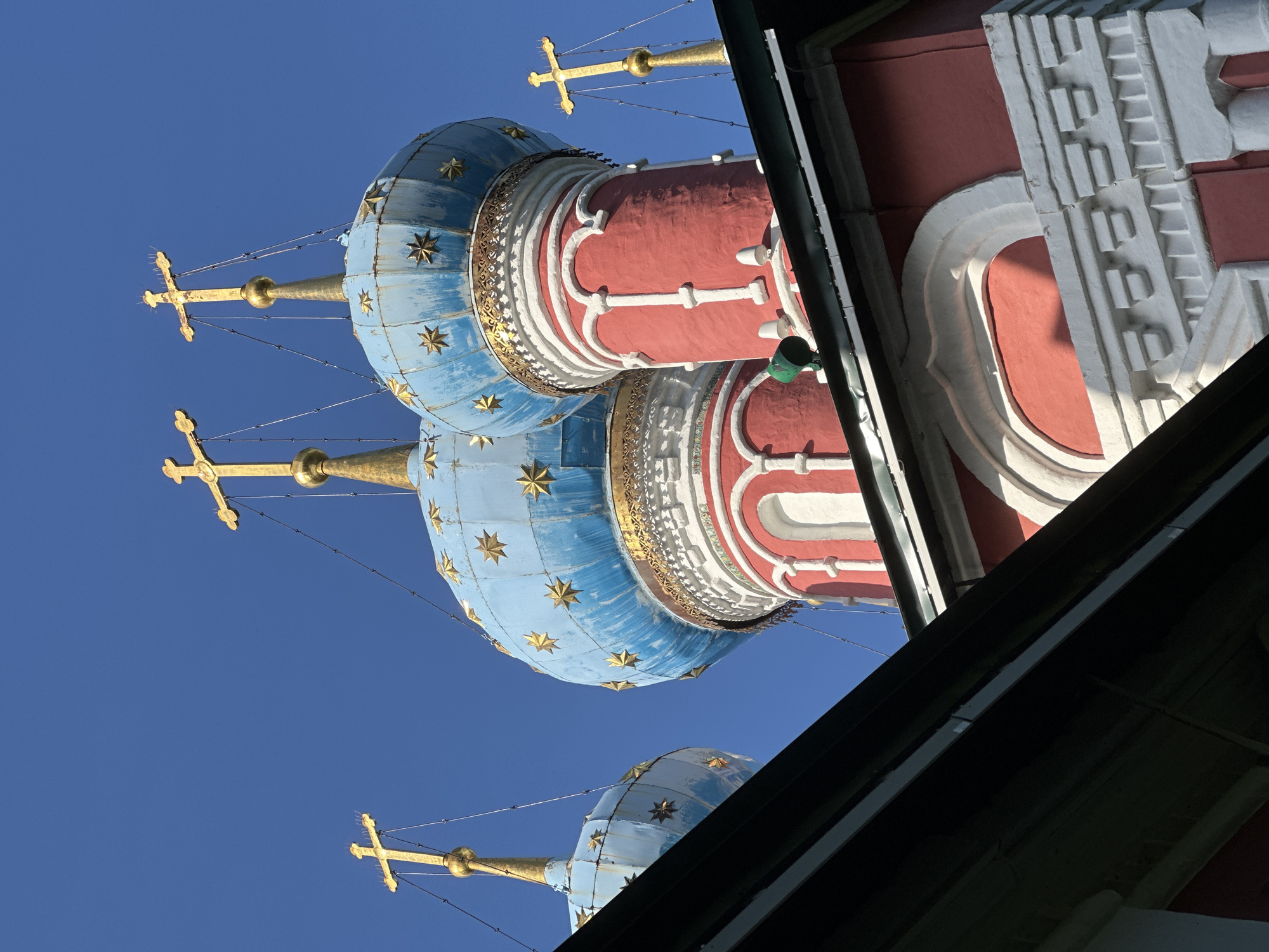 The image shows a close-up, low-angle view of the upper portions of multiple onion domes of a Russian Orthodox church, set against a clear, bright blue sky. The domes are predominantly blue with gold stars, topped with gold crosses. The lower sections of the domes are red with white decorative trim. A dark, angled roofline, possibly of another building or an overhang, partially obscures the bottom left of the frame. The architectural style suggests a traditional Russian church. The city of Terpilovo, Russia, is mentioned as the location. There are no people visible in the image.