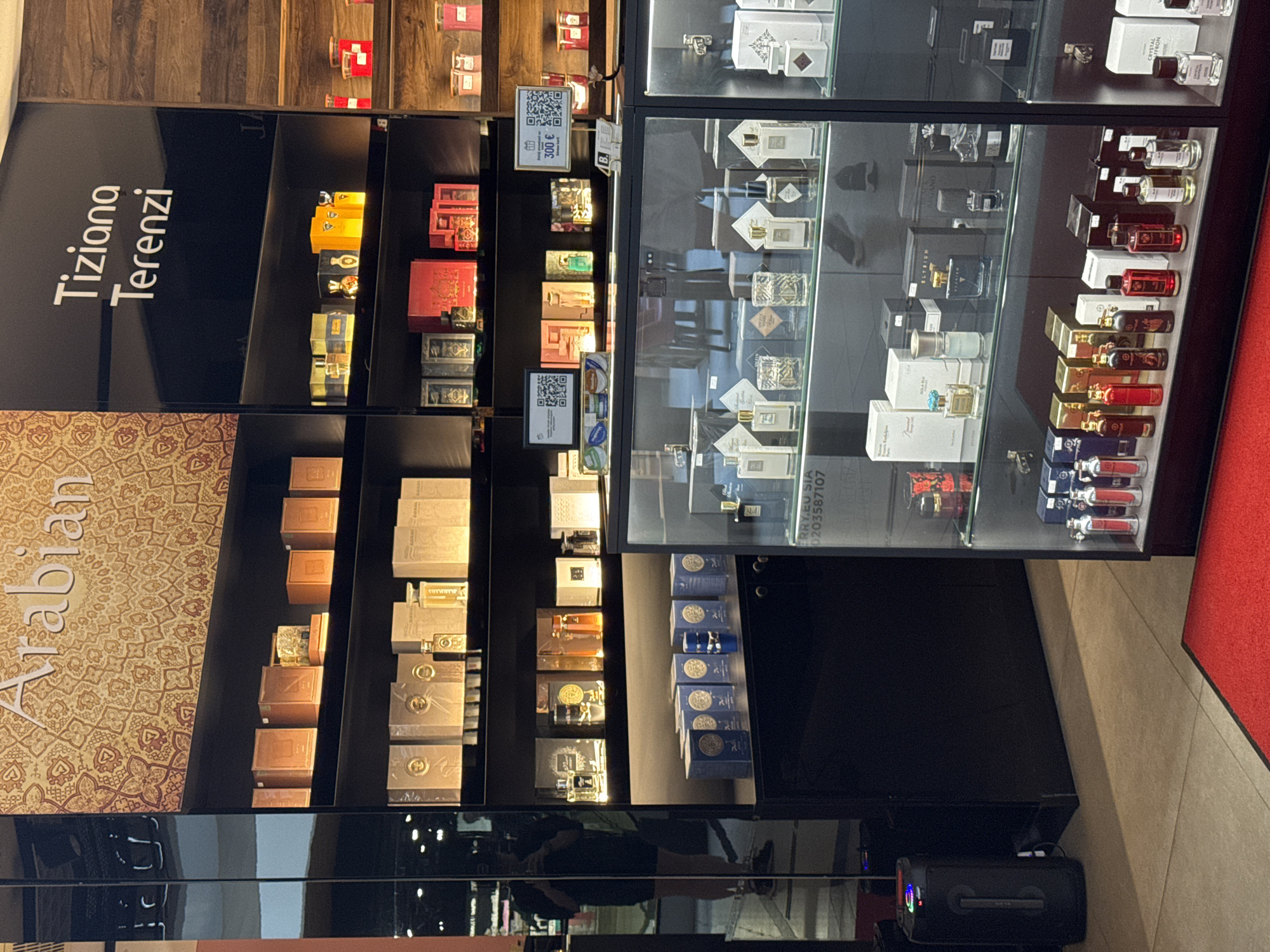 The image presents an interior view of a luxury fragrance store, likely located in Riga, Latvia, given the provided context. The setting is modern and sophisticated, featuring dark display shelves and illuminated glass cabinets showcasing an extensive collection of perfumes.
On the left, a prominent black panel displays the brand name "Tiziana Terenzi" in white, vertical text. Below this, and continuing downwards, are dark shelves holding numerous packaged products, predominantly perfume boxes in various designs and colors, including gold, red, and silver. A patterned wall section, rich with intricate designs, features the word "Arabian" in stylized white text, indicating a dedicated section for Arabian or Middle Eastern-inspired fragrances. Small signs with QR codes are affixed to the shelves; one clearly indicates a price of "300 €". Other visible text on packaging includes "MARMARA".
To the right, two tall, glass-fronted display cases are packed with individual perfume bottles and their accompanying boxes. The bottles come in diverse shapes, sizes, and colors, ranging from clear glass to opaque and dark designs. Brands such as "CRYSTAL SAFFRON" are discernible on some bottles and boxes. Reflections on the glass suggest the presence of a person (likely the photographer) and other elements of the store's interior.
The floor on the right features a vibrant red carpet, contrasting with what appears to be beige or light-colored floor tiles further left. At the bottom of the image, a black, glossy surface reflects the upper displays, and a black portable speaker or similar electronic device rests on the floor. Small text "FRRY.EU SIA 0203587107" is also visible. The artificial lighting within the store highlights the products, creating an inviting retail environment.