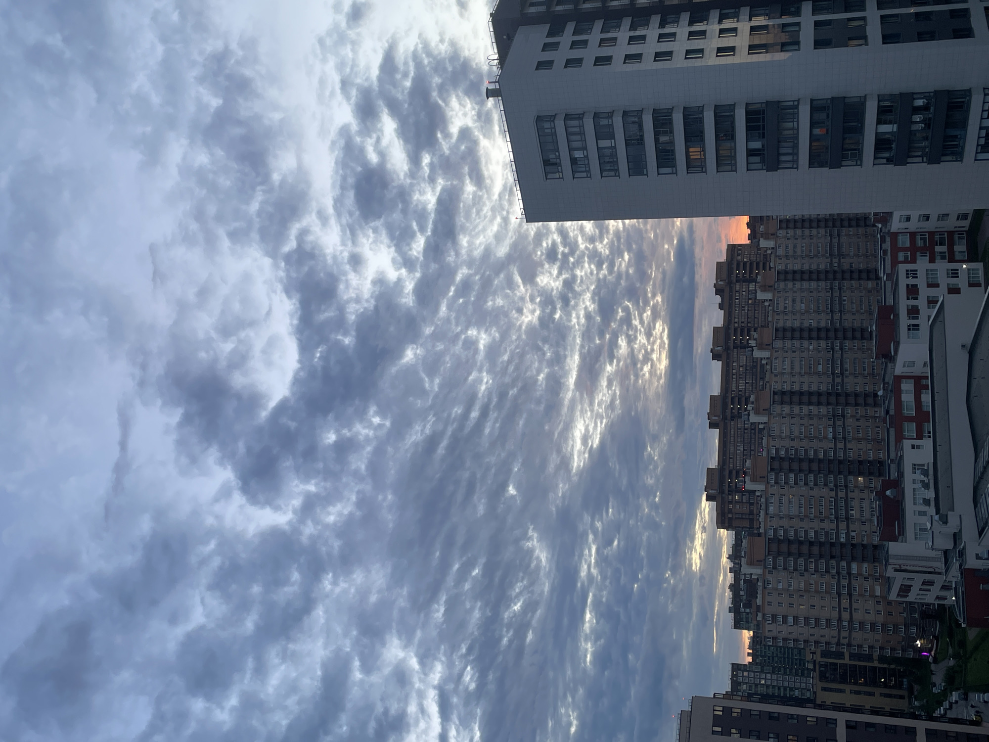 This is a vertical, eye-level, medium shot of a city skyline in Sinyavino, Russia, at dusk. The sky is filled with dramatic, textured clouds that are dark gray and blue, with streaks of lighter gray and hints of orange and yellow from the setting sun peeking through. In the foreground, on the right, is a tall, modern apartment building with a white facade and dark windows. To the left and in the background are numerous other apartment buildings of varying heights and architectural styles, with many windows visible. Some of the buildings are illuminated, suggesting people are home. The overall impression is of a dense urban environment under a dynamic, cloudy sky. No people are visible in the image.
