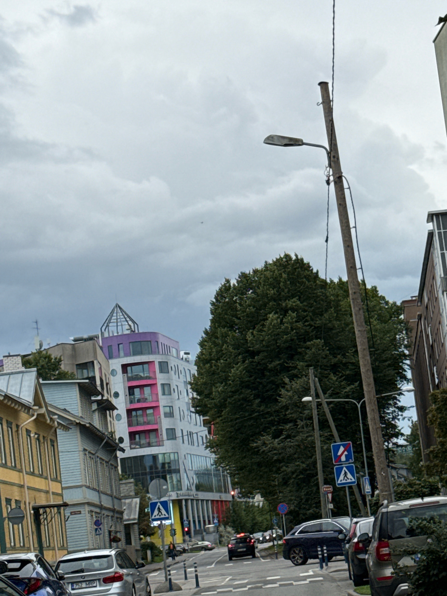 The image depicts a street scene in Tallinn, Estonia, under an overcast sky. The setting appears to be a city street with a mix of older, colorful buildings and modern architecture. In the foreground, several cars are parked or driving on the road, including a silver BMW with a license plate partially visible. A prominent wooden utility pole with a streetlight stands to the right of the frame, leaning slightly.
Behind the cars, a row of traditional wooden houses painted in yellow and blue line the street, suggesting a historic district. Further down the street, a striking modern building features a colorful facade with sections in pink and purple, topped with a glass-domed structure. Dense green trees are visible, providing a contrast to the urban environment.
Traffic signs, including a pedestrian crossing sign and what appears to be a road closure sign, are present along the street. A few people can be seen walking on the sidewalk in the distance. The overall mood is subdued, likely due to the cloudy weather. The time of day appears to be daytime, but the cloudy conditions create a dim, diffused light. There are no strong indicators of a specific event taking place, suggesting a typical day in the city.