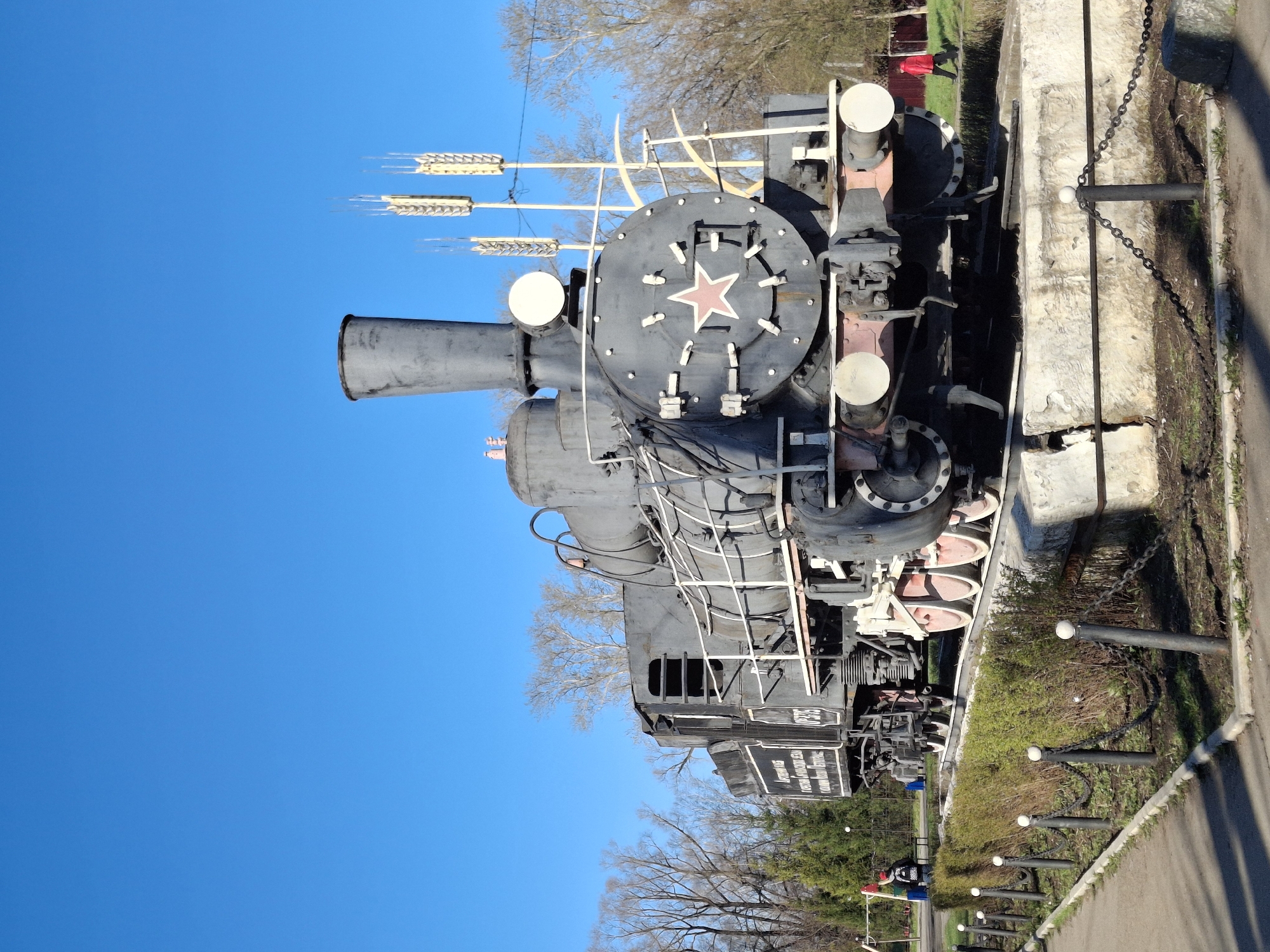 A vintage, dark gray steam locomotive, identified by the number "Л-3235" (L-3235), is prominently displayed outdoors as a static monument. The locomotive faces to the left within the frame, set against a bright, clear blue sky.
The front of the engine features a large, circular plate adorned with a striking red five-pointed star, a common symbol of the Soviet era. Above the star, several gold-colored, spike-like elements, possibly symbolizing wheat sheaves, extend upwards. The locomotive's large smokestack, boiler, and various intricate mechanical components are clearly visible. The driving wheels, with their distinctive red and white accents, are partially shown beneath the engine's body.
The tender, the section immediately behind the main engine, bears white Cyrillic text on a black background. The text "Л-3235" is clearly legible, along with the inscription "ПОСТРОЕН НА КОЛОМЕНСКОМ ЗАВОДЕ В 1953 ГОДУ" (BUILT AT THE KOLOMNA PLANT IN 1953 YEAR). Additional lines of text, though somewhat obscured, likely elaborate on the locomotive's historical significance or achievements.
The setting is an outdoor area in Sarapul, Russia, likely a park or public square where the locomotive is preserved. It rests on tracks next to a barrier composed of concrete blocks and metal posts connected by chains. A paved path runs parallel to the barrier. The background reveals bare deciduous trees, suggesting a season of late winter or early spring, alongside patches of green grass and bushes. The scene is brightly lit by the sun, casting distinct shadows. In the far distance, indistinct figures and colorful structures, possibly part of a playground, are faintly visible, indicating a public recreational space. No specific activity is depicted, and the overall atmosphere is calm and observational.