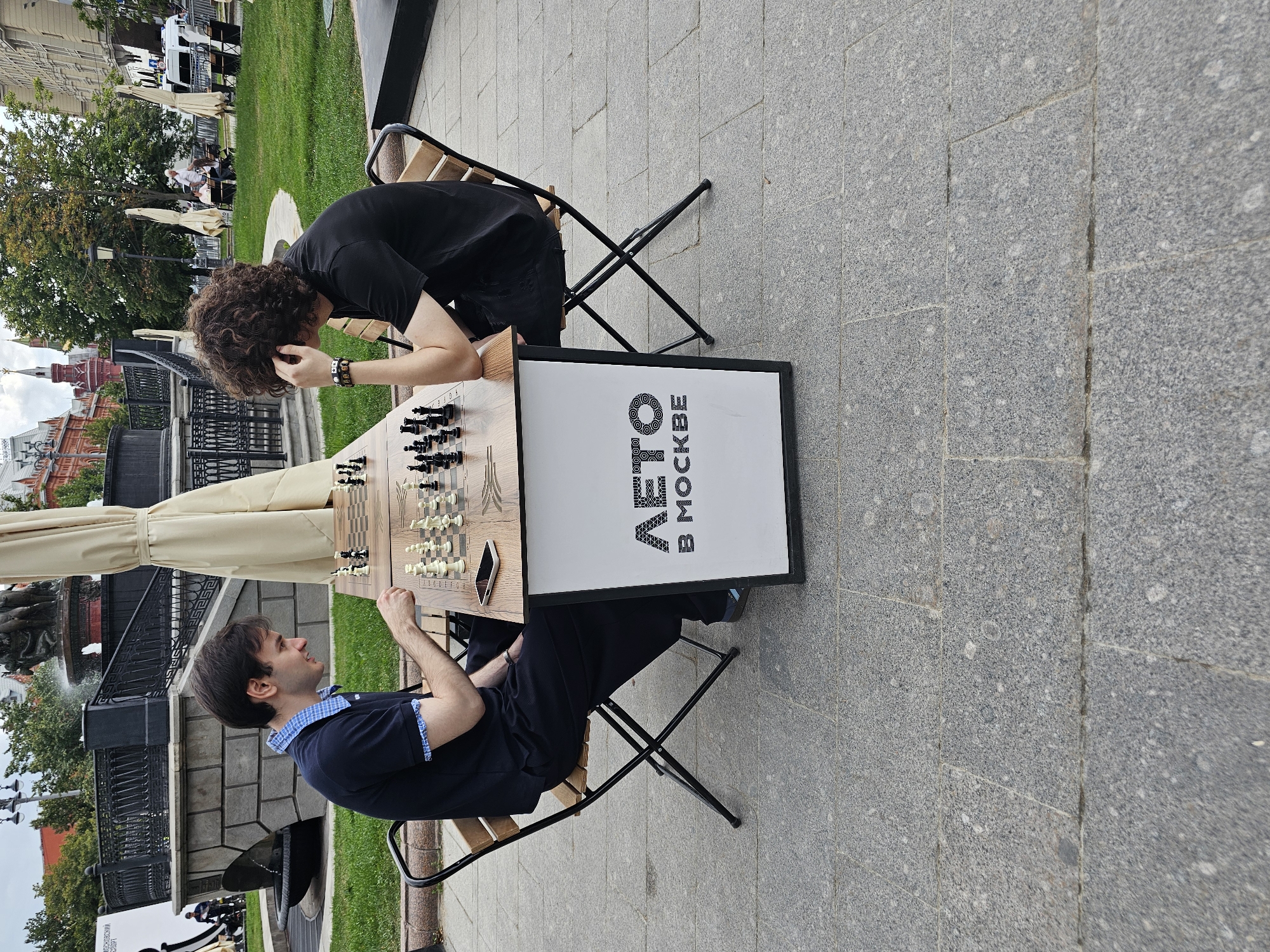 Two men are playing chess at a table set up outdoors. The man on the left, wearing a dark blue polo shirt and dark pants, is looking at the chessboard. The man on the right, with curly brown hair and wearing a black t-shirt, has his hand on his forehead, appearing to be thinking. They are seated on folding chairs at a table with a chessboard and pieces. A sign on the front of the table reads "АВТО В МОСКВЕ" (Cars in Moscow). The background shows a park-like setting with trees, a fountain, buildings in the distance, and other people sitting on benches. The scene appears to be in a public square in Moscow, Russia, likely near historical architecture given the style of the buildings. The pavement is made of granite slabs.