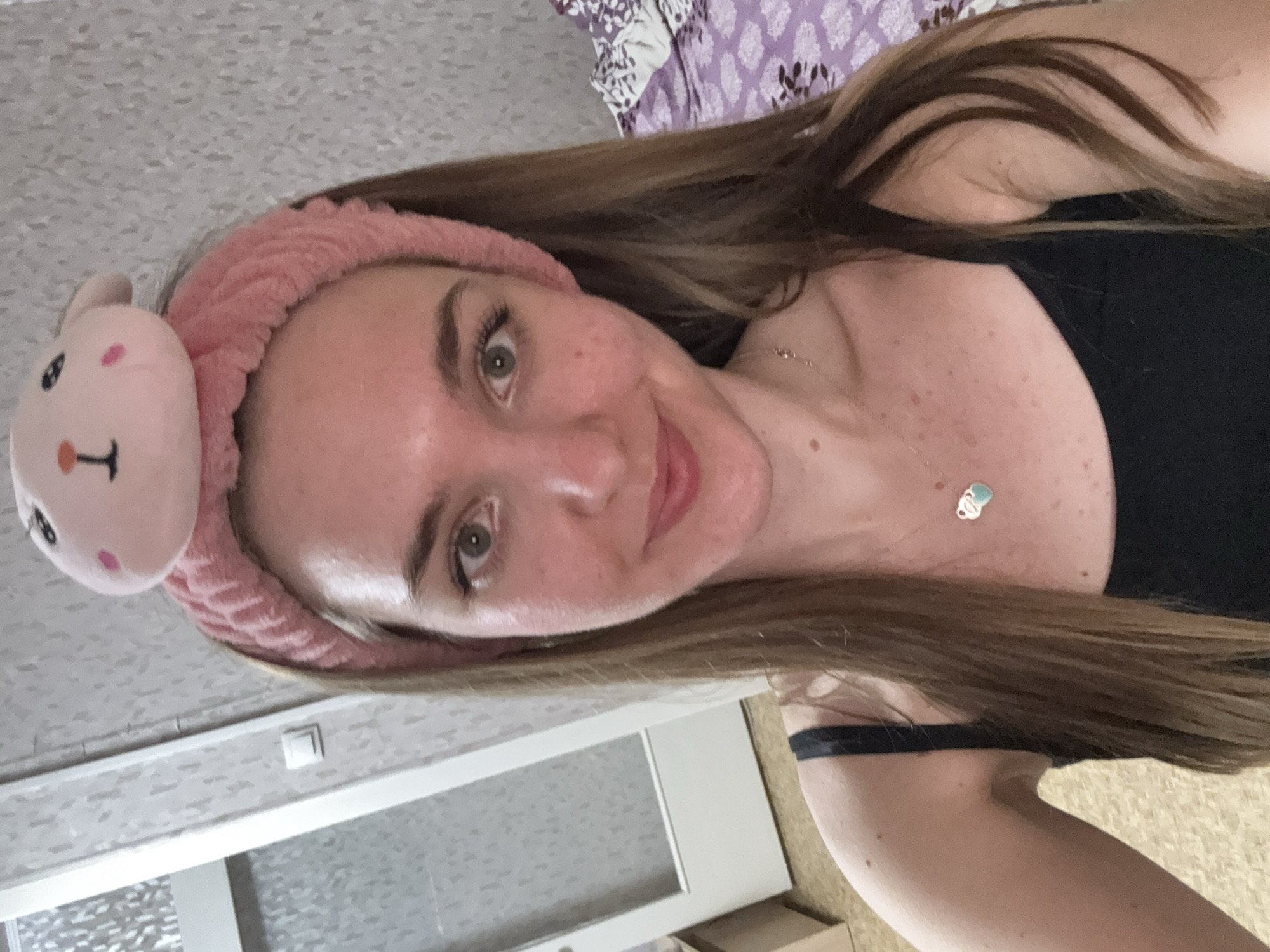 The image shows a woman with light brown hair, a pink bunny-ear headband, and a black top. She is wearing a delicate necklace with a light blue pendant. The background includes a textured wall and a partially visible patterned bedspread. A white door with a frosted glass panel is also visible, along with a section of the floor. The location is assumed to be Perm, Russia, but no elements in the photo confirm this, it is only speculative.