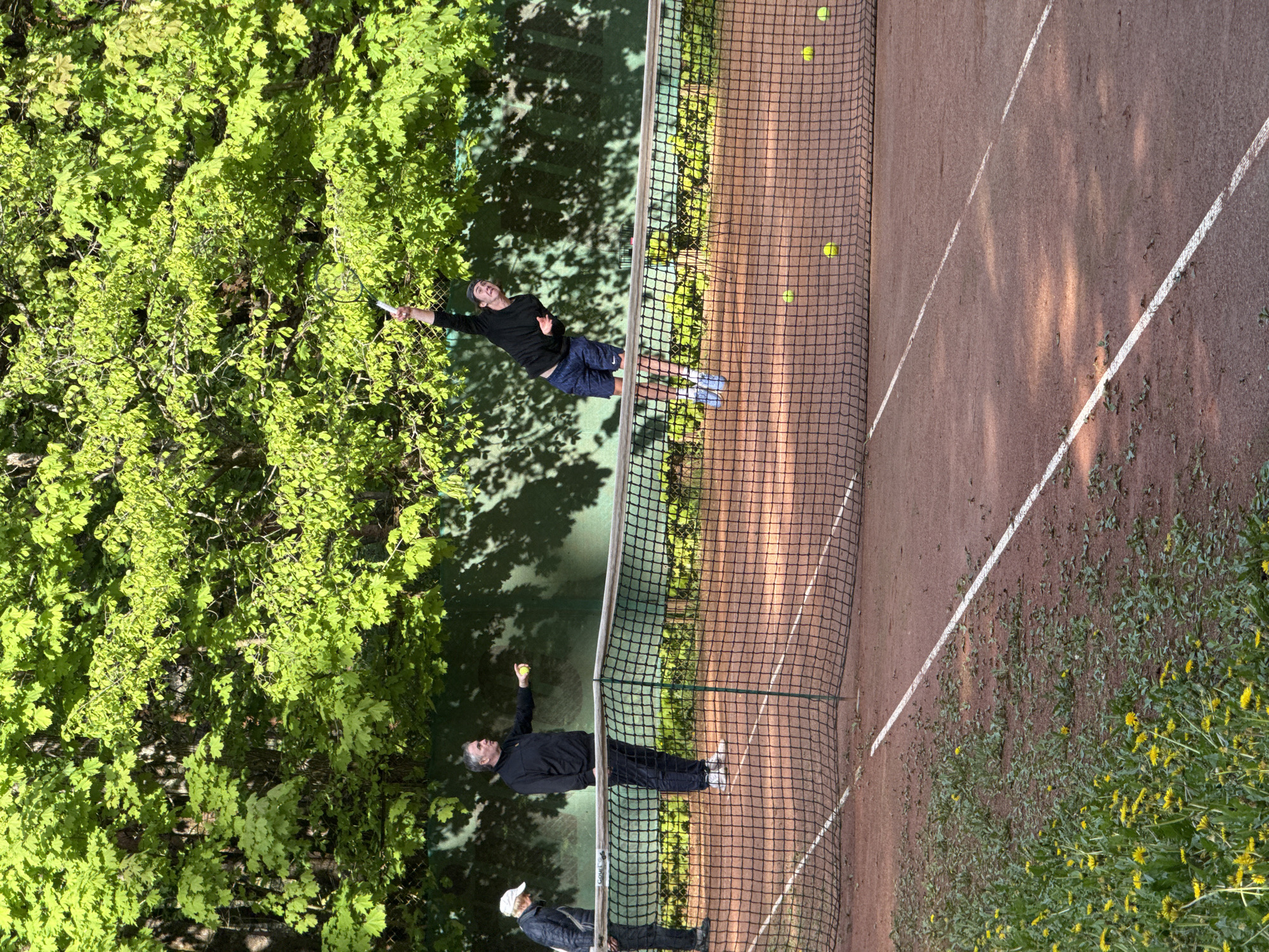 The image shows a tennis court, possibly in the city of Unknown, Russia. The court is made of clay and has a net in the middle. On one side of the net, a man wearing a black shirt and blue shorts is serving a tennis ball with a racket. On the other side of the net, another man in a black jacket is holding a tennis ball. A third person, wearing a white cap and a dark jacket, is standing near the second man. Several tennis balls are scattered on the court. The background consists of green trees and some yellow flowers. The picture appears to be taken during the day, with natural light.