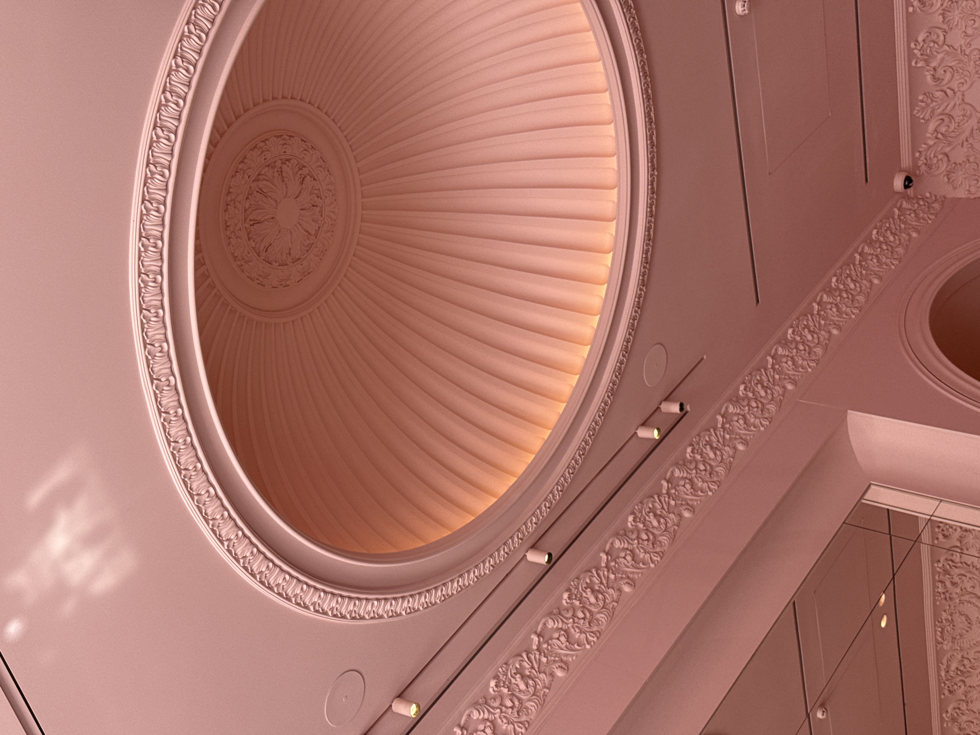 The image shows a decorative pink ceiling with an oval-shaped dome. The dome has a fluted design and a central rosette. Ornate molding with floral motifs frames the dome and runs along the edges of the ceiling. Several spotlights are visible, as are what appear to be air vents and security cameras. The overall aesthetic suggests an interior space with classical architectural influences. The image does not provide any information about the location in Kyiv, Ukraine.