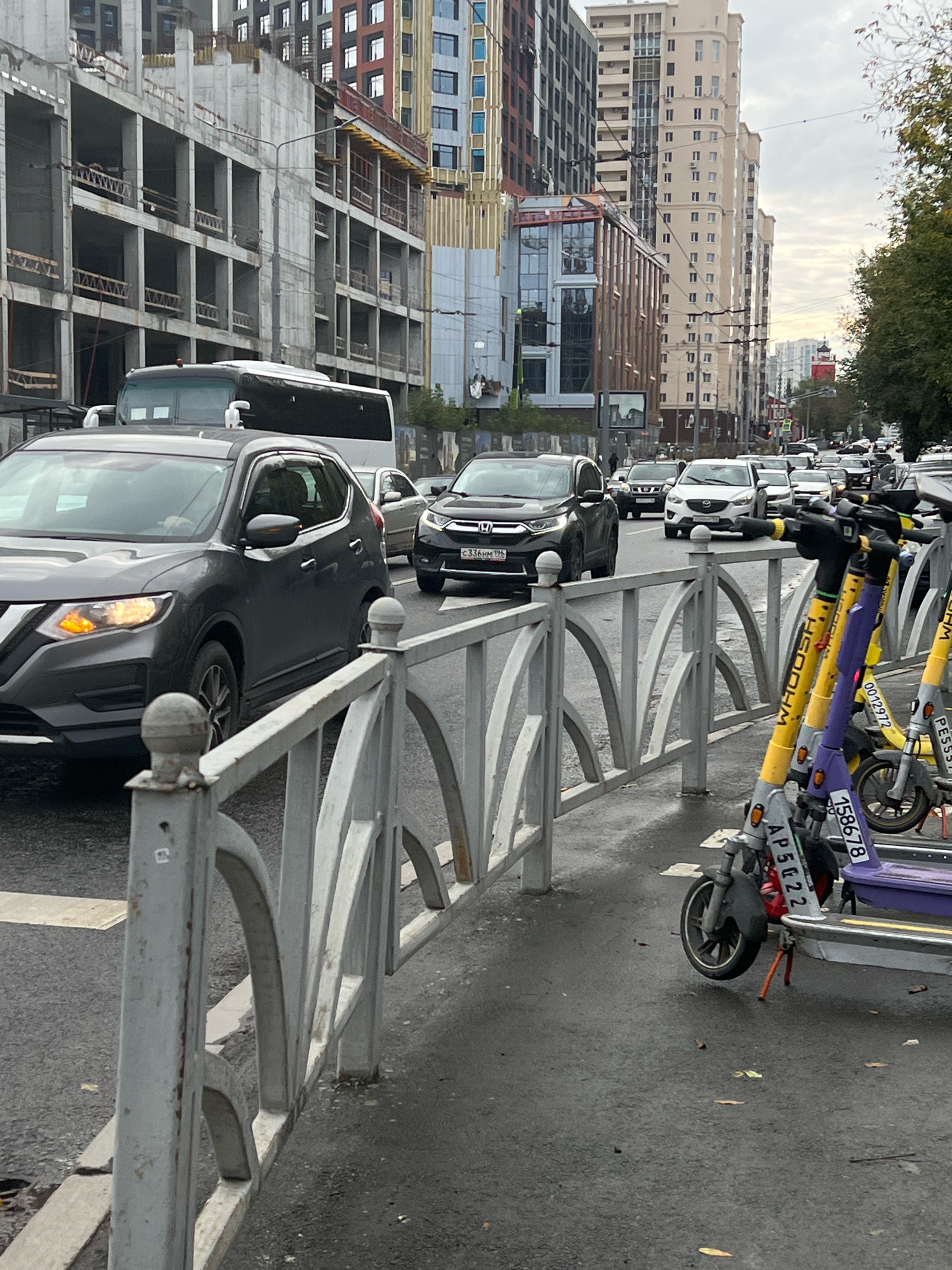 The image depicts a street scene in Yekaterinburg, Russia, likely during daytime given the illumination. In the foreground, several electric scooters are parked along a sidewalk separated by a metal fence. The scooters are colored yellow and purple and bear various identification numbers and markings, including "WHOOSH," "0012972," "PESSI," "AP5G22," and "158678."
Behind the fence, a gray Nissan X-Trail SUV is stopped in traffic, its front right turn signal illuminated. Next to it, a black Honda CR-V is also in traffic, with its license plate visible as "C 336HM 196." Other vehicles, including a white SUV and a bus, are visible further in the traffic flow.
The background features a mix of urban architecture. To the left, a multi-story building under construction with a concrete facade and exposed structural elements is prominent. To the right and further back, several tall, modern apartment buildings with numerous windows and balconies rise into the sky. The overall environment suggests a busy city street with ongoing development. The pavement appears wet, indicating recent rain or damp conditions.