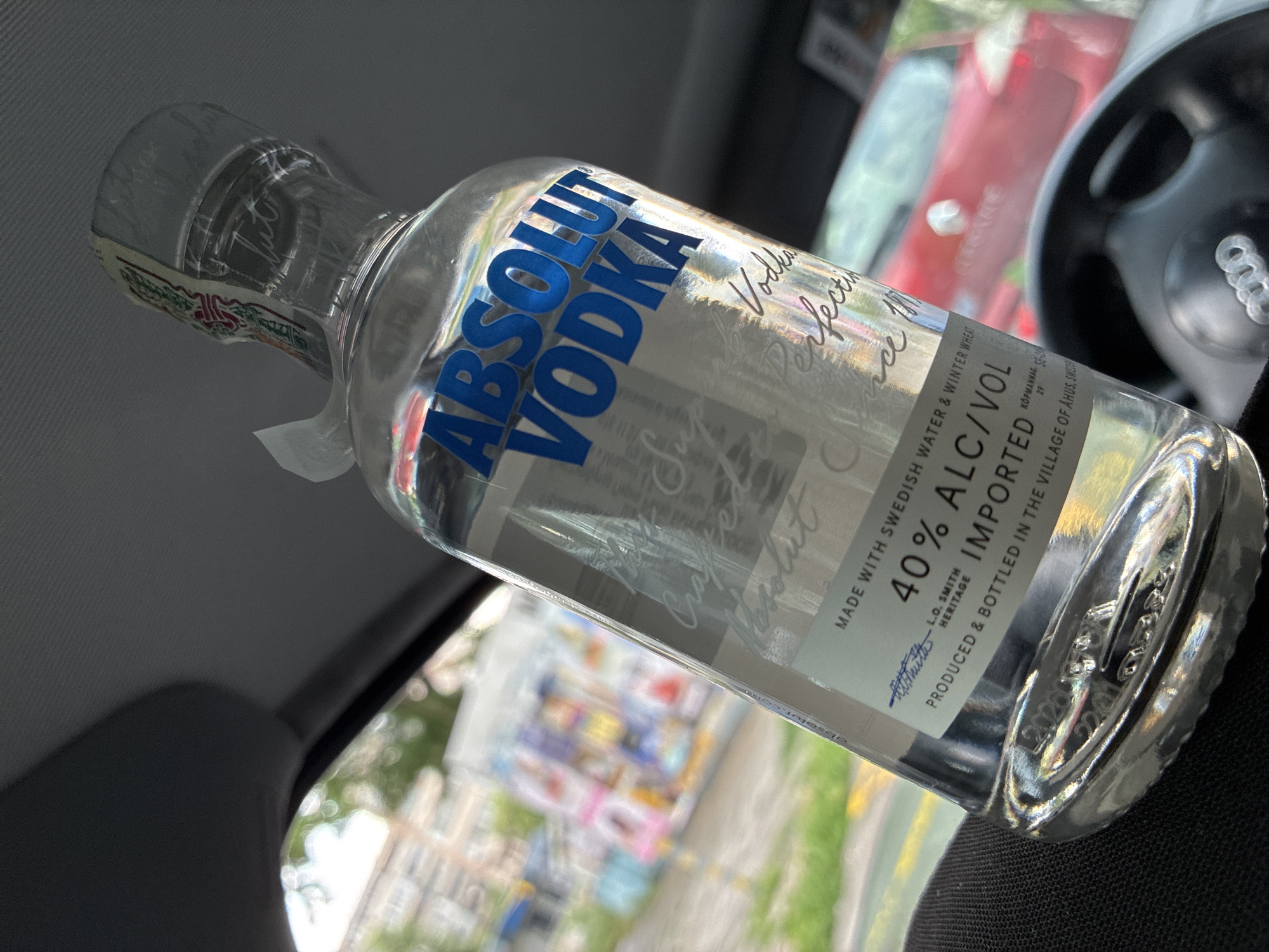 A bottle of Absolut Vodka lies on the front passenger seat of a car. The bottle is mostly full, with the brand name prominently displayed in blue lettering. Text on the label includes "Made with Swedish Water & Winter Wheat" and "40% Alc/Vol". The car's steering wheel, with the Audi logo, is visible in the background. The scene is viewed through the car window, showing a blurred cityscape with buildings and greenery. The image does not offer sufficient information to determine the exact location in Varna, Bulgaria or definitively show it is in Varna, Bulgaria.