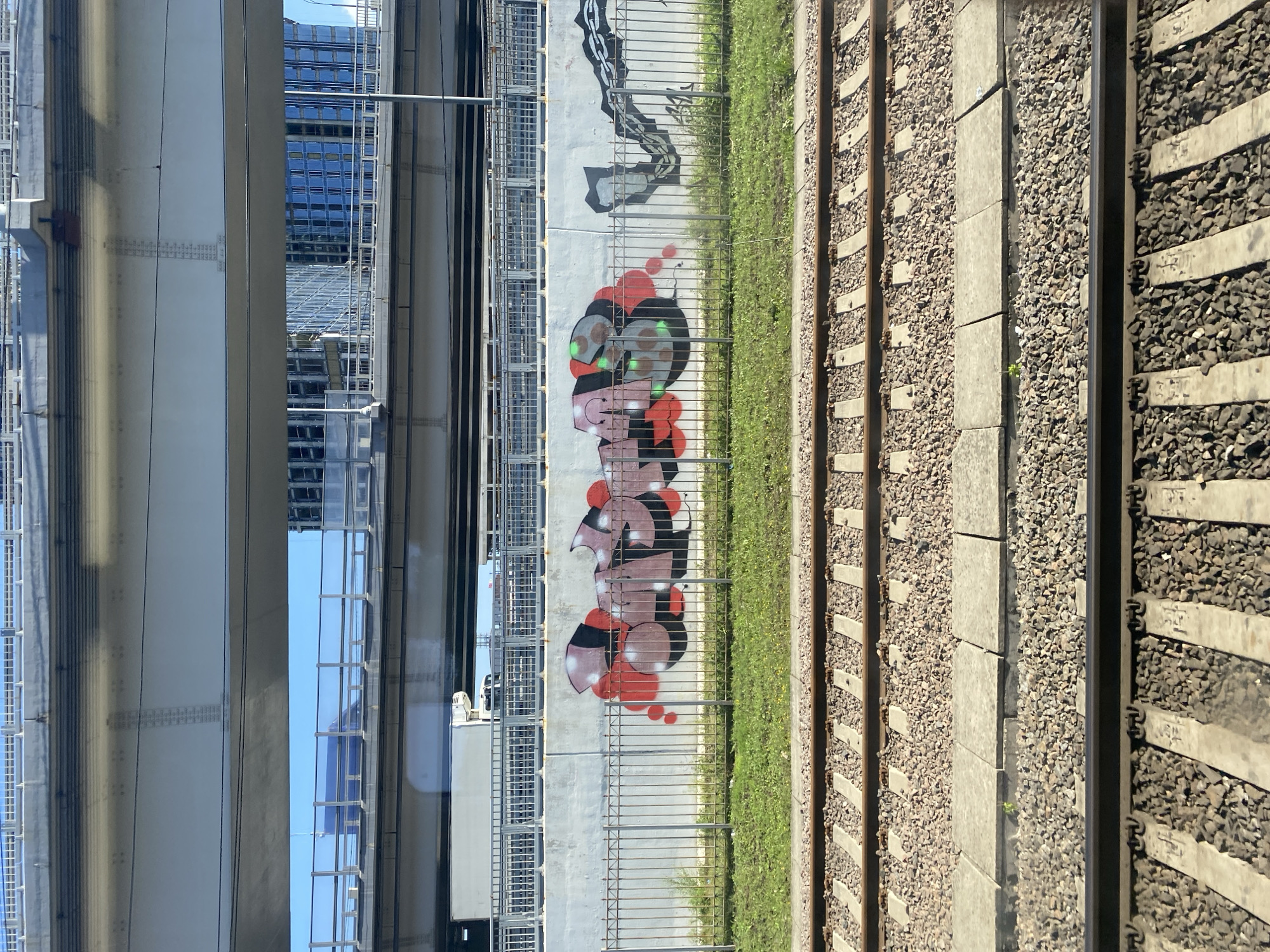 The image captures a scene from a moving vehicle, likely a train, looking out at an urban environment. In the foreground, railroad tracks are visible, laid on ballast. Beyond the tracks, a low green grassy area is separated from a concrete wall by a metal fence. The concrete wall features graffiti in vibrant colors, primarily shades of red, pink, black, and green, forming abstract shapes and lettering. To the right of the graffiti, a stylized chain is also spray-painted on the wall. In the background, multiple overpasses and bridges dominate the view, with modern glass-fronted buildings peeking through the gaps. The sky is a clear blue. The overall impression is one of an urban landscape with elements of infrastructure and street art.