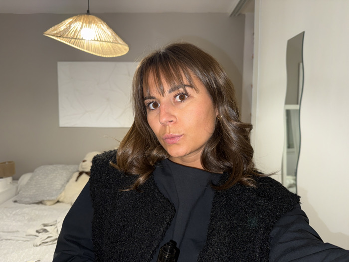 A woman with brown hair and bangs poses indoors, wearing a black top and a textured black vest. Behind her is a bed with neutral-colored pillows and a teddy bear. A woven pendant light hangs from the ceiling above a white abstract art piece. A wavy mirror stands against the wall beside a white closet.