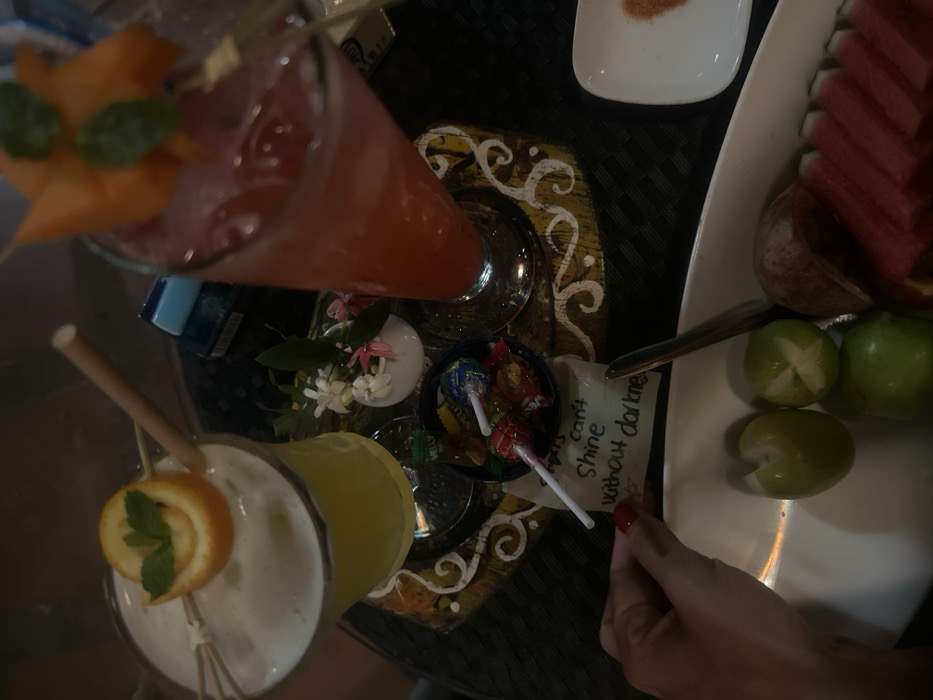 The image displays a high-angle view of a table setting within a dimly lit establishment, likely in Thuận Hóa Ward, Vietnam. Two garnished cocktails are positioned on a round, dark brown tray featuring decorative gold and white swirl patterns. The cocktail on the left is a yellow, frothy drink in a stemmed glass, garnished with a spiral orange peel and mint leaves, accompanied by a bamboo straw and two wooden skewers. The cocktail on the right is a red drink with ice, served in a tall stemmed glass, garnished with a piece of carved orange-colored fruit (possibly starfruit or papaya) and mint leaves, also with a bamboo straw.
Between the cocktails, on the decorative tray, rests a small white vase containing small white and pink flowers with green foliage. Adjacent to the flowers, a small blue bowl holds an assortment of colorful lollipops and candies, some with visible wrappers (one reading "MELLY").
Below the tray, a white rectangular dish contains an arrangement of cut fruits: several green plums or limes (some scored), multiple slices of watermelon, and slices of a reddish fruit, possibly apple or plum. A metallic utensil, appearing to be a spoon, is visible beside the fruit. To the right of the fruit dish, a small white square dish contains a reddish-orange powder, likely a spice mix or chili salt.
A person's hand, with red painted fingernails, is visible in the lower-left foreground, holding a small, light-colored note. The handwritten text on the note reads: "Stars can't Shine without darkness." In the background, partially obscured, is a blue object, possibly a cigarette pack, with some text visible. The table surface beneath the items is dark with a textured or patterned fabric.