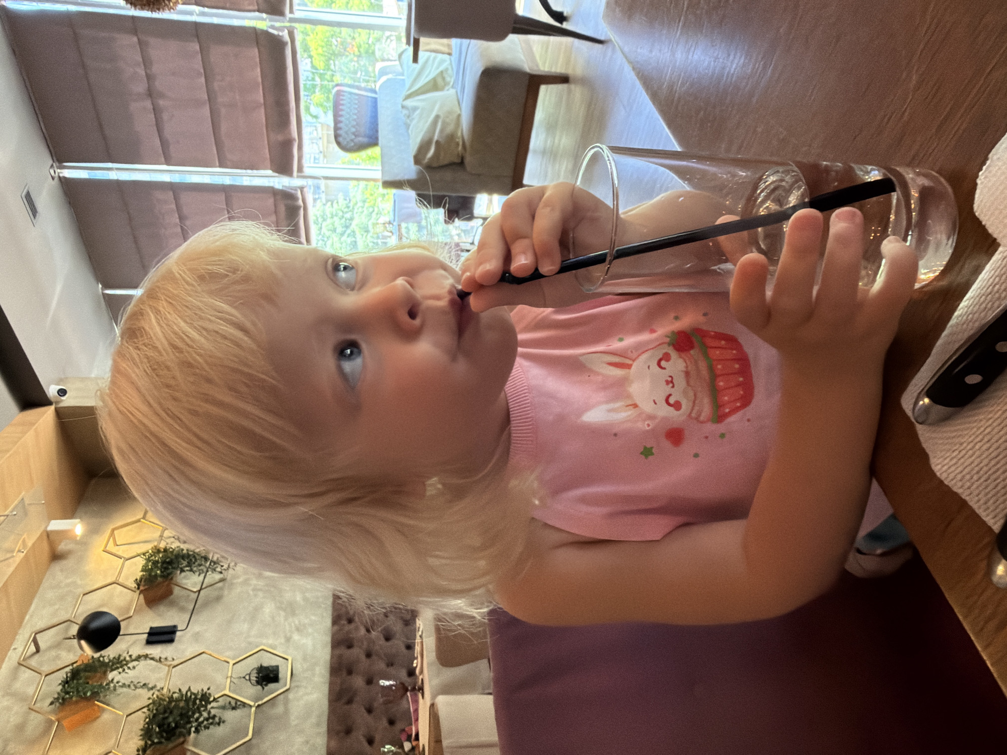 A young girl with blonde hair is drinking from a glass with a straw. She is wearing a pink t-shirt with a cupcake and bunny design. She is sitting at a table in what appears to be a restaurant or cafe. In the background, there are windows with blinds, and some upholstered chairs and a sofa. There is also a decorative wall feature with geometric shapes and plants. The lighting suggests it could be daytime.