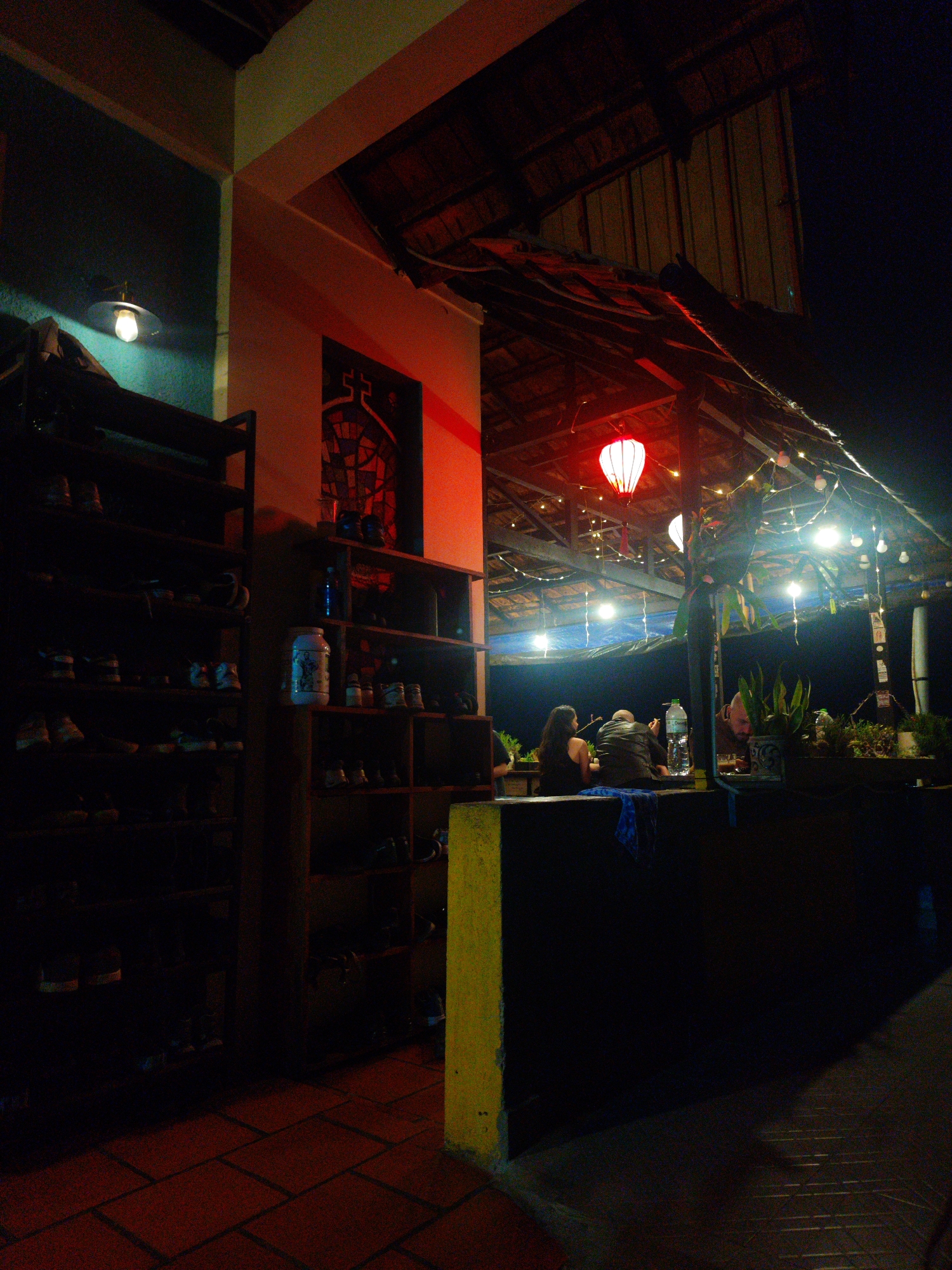 The photograph depicts an evening scene at a building in Da Lat, Vietnam. A red lantern hangs beneath a thatched roof extension, illuminating a group of people sitting at a counter. String lights are strung across the roof. Inside the building, two wooden shelving units stand against the wall, one blue, the other white, holding miscellaneous items including shoes and jars. The floor is tiled with red bricks. The overall lighting is low, creating a dim atmosphere.
