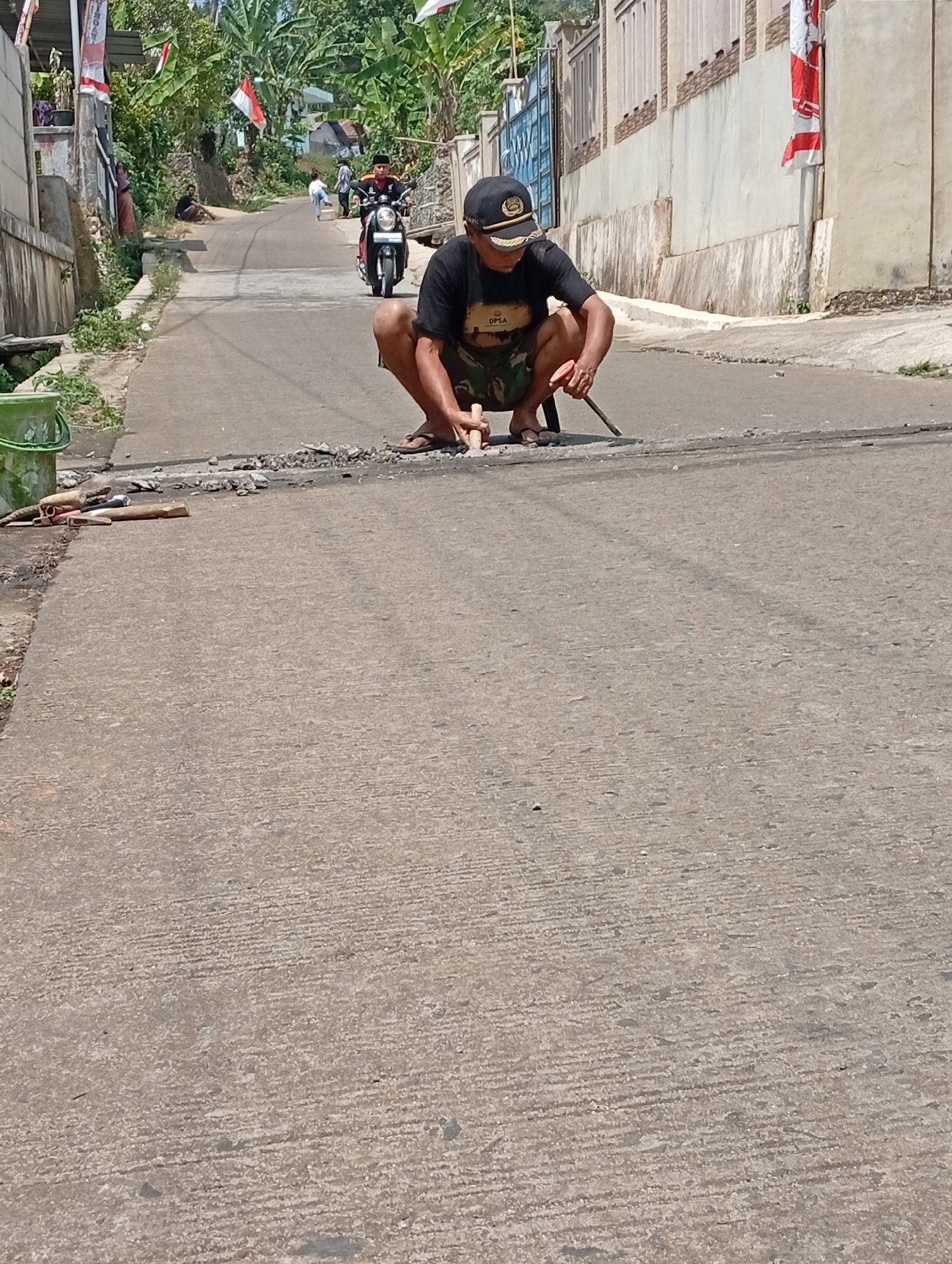 In Cisitu, Indonesia, a man is crouched on a paved road, holding a pointed tool and appearing to chip away at the surface. He wears a dark t-shirt with a logo, camouflage shorts, a baseball cap, and sandals. In the background, a motorcycle with a rider passes by, and other individuals are visible further up the road. The environment suggests a sunny day with houses and greenery lining the street.