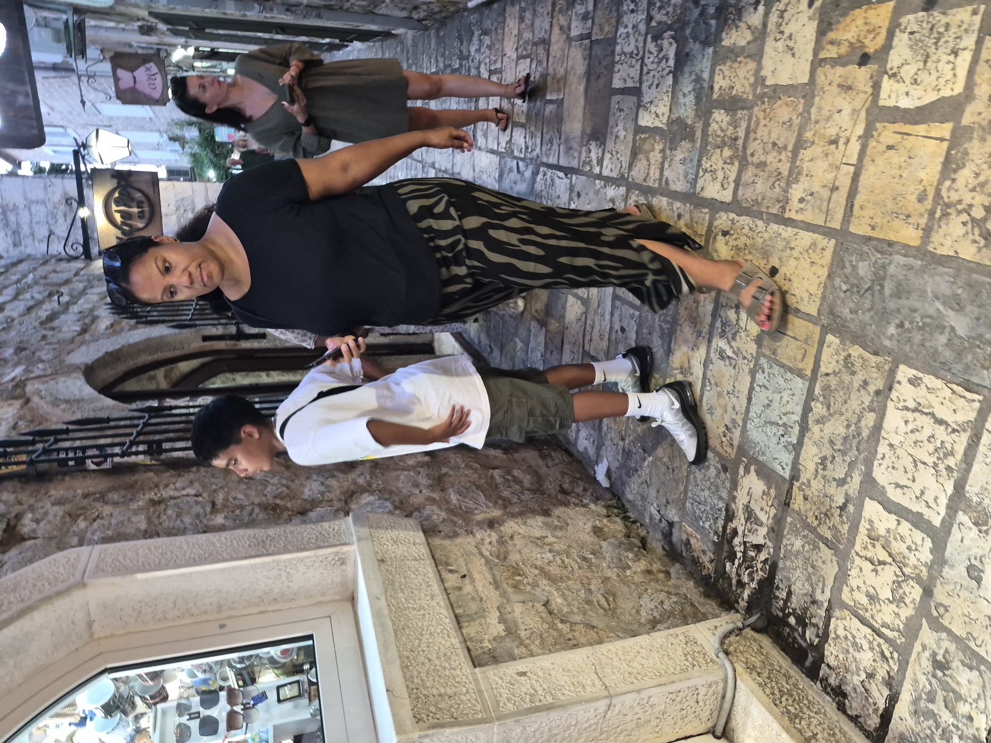 In Budva, Montenegro, on a cobblestone street lined with stone buildings, a young boy stands in front of a shop window, looking at the merchandise. He is wearing a white t-shirt and khaki shorts. Beside him, a woman in a black t-shirt and patterned pants walks past, holding a phone. Further down the street, another woman in a green dress is also looking at her phone. The scene suggests a casual stroll through a historic European town, possibly during a holiday. The lighting is dim, indicating it could be evening or a cloudy day.