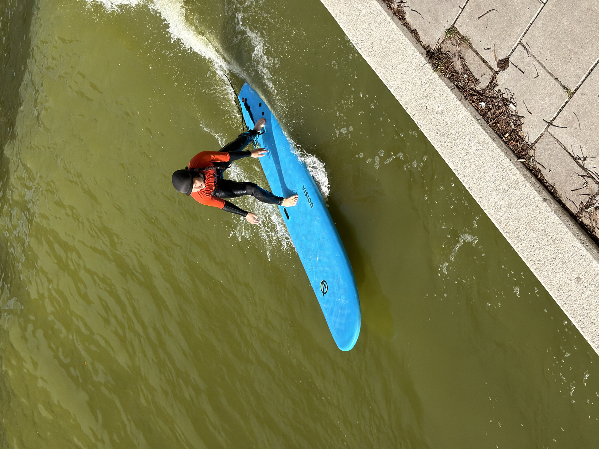 An individual in a black wetsuit with an orange long-sleeved top and a black helmet is surfing on a blue surfboard on a green, wavy body of water. The surfboard has the word "VISION" printed on it and a black circular logo. The person is standing on the board with their arms outstretched for balance. The water is rippling and shows signs of movement, with white foam trailing behind the surfboard. To the right of the water, there is a concrete embankment or walkway, and beyond that, a paved area with some debris, including twigs and dry leaves. The scene appears to be an urban environment, possibly a canal or river within the city of Rotterdam, Netherlands, given the architectural style of the embankment and the presence of urban waters.
