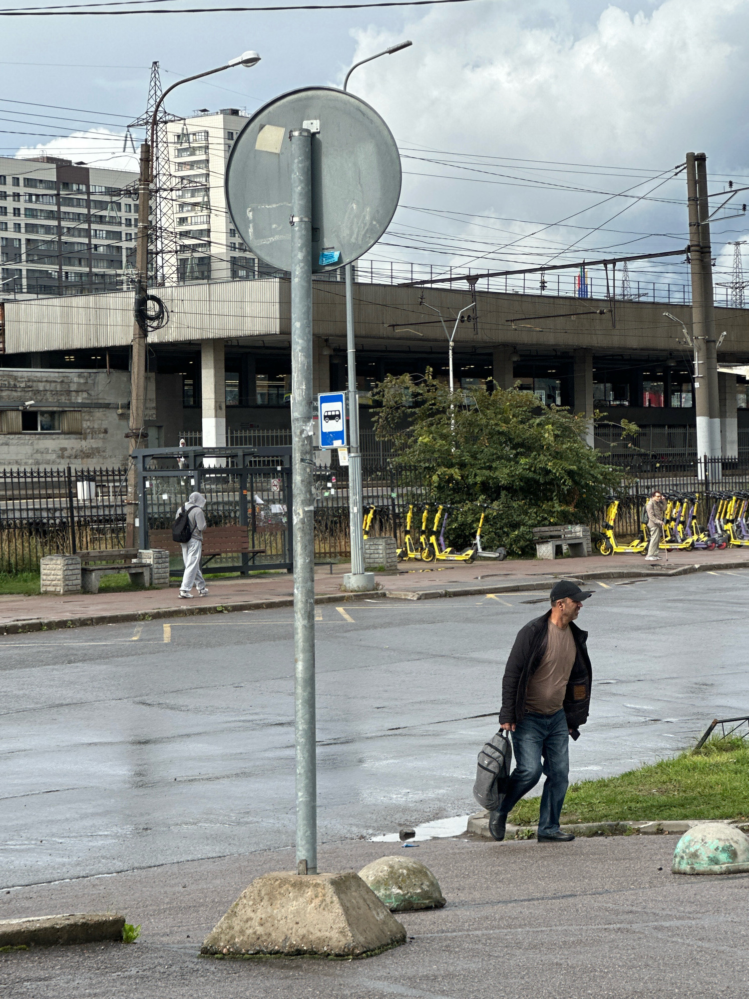 A man wearing a black jacket, brown t-shirt, and blue jeans walks on a wet asphalt road, carrying a gray backpack in his right hand. He is walking away from the viewer and towards the right side of the frame. In the background, a person in a grey hooded sweatshirt and light-colored pants walks on a sidewalk. Behind them is a bus stop shelter with a blue bus sign. Further back, a row of yellow and purple electric scooters is parked. The scene appears to be an outdoor urban environment, likely in Murino, Russia, with buildings, power lines, and street infrastructure visible. The sky is overcast.