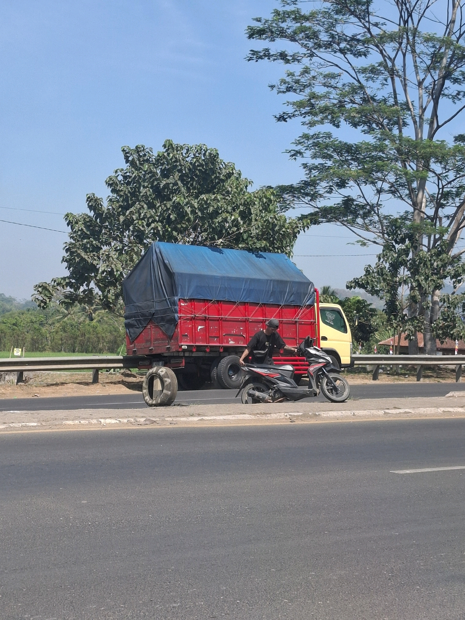 A person in a black t-shirt and cap is standing next to a parked red truck with a blue tarp covering its cargo. The truck is on the side of a road, and a black motorcycle is parked beside it. The truck has a spare tire leaning against its rear, and another tire is visible on the truck itself. The scene appears to be in Babakan, Indonesia, with trees and a clear blue sky in the background.