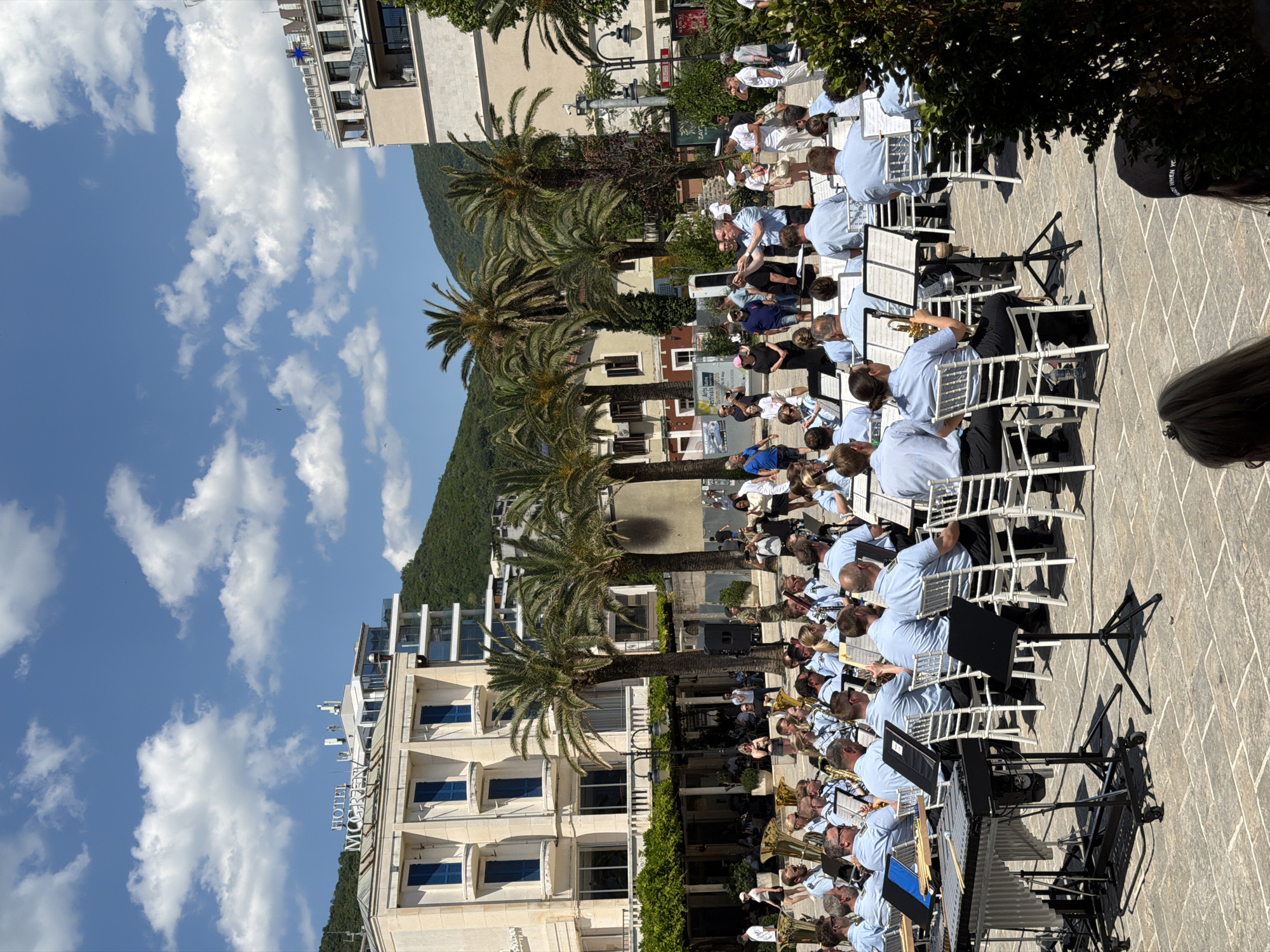 A wide-angle medium shot depicts a musical ensemble performing in an outdoor setting. The performers, dressed in light blue shirts and dark pants, are seated on white chairs with music stands in front of them, indicating their instruments.
Palm trees are visible between the building and the ensemble. The buildings are multi-storied with light-colored facades and blue shutters. A hillside rises in the background. The weather is sunny, with a blue sky and scattered clouds.
The scene appears to be in or near the city of Boreti, Montenegro, given the architecture, vegetation, and general setting.