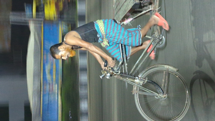 A man rides a bicycle on a road, with bare feet and wearing a black tank top and a blue and green striped wrap-around skirt. The bike is old and rusty, with a large front wheel and a smaller back wheel. The man's beard is orange. The background is blurred, but it appears to be a busy street. The image is slightly tilted, giving a sense of movement.