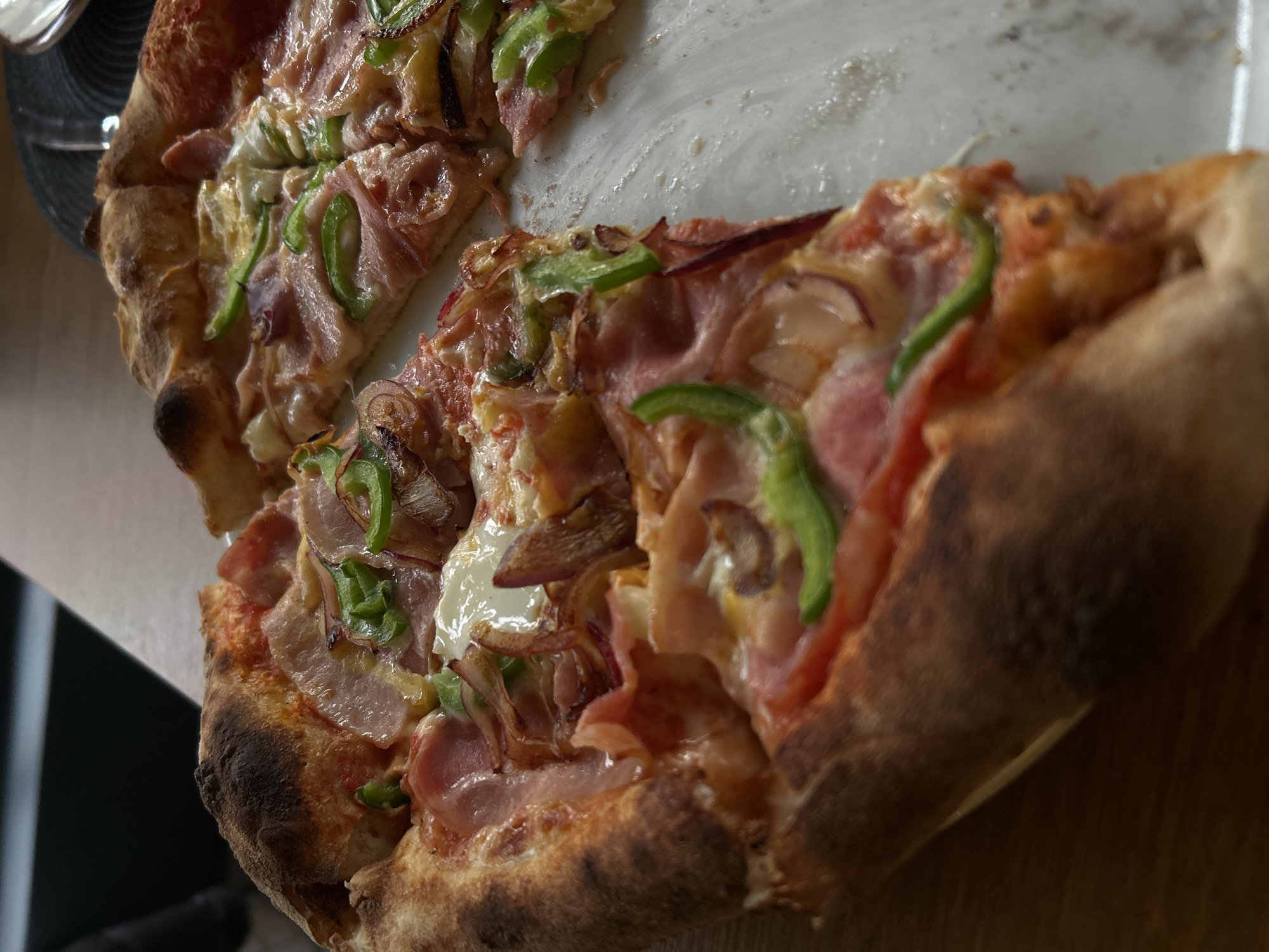 The image shows a partially eaten pizza with a charred crust, toppings of prosciutto, sliced green bell peppers, and red onions on a light-colored plate set on a wooden surface. A black plate is visible in the background on the left. The pizza appears to be freshly made. The location is unknown, but is described in the prompt as the city of Sofia, Bulgaria. No people are visible.