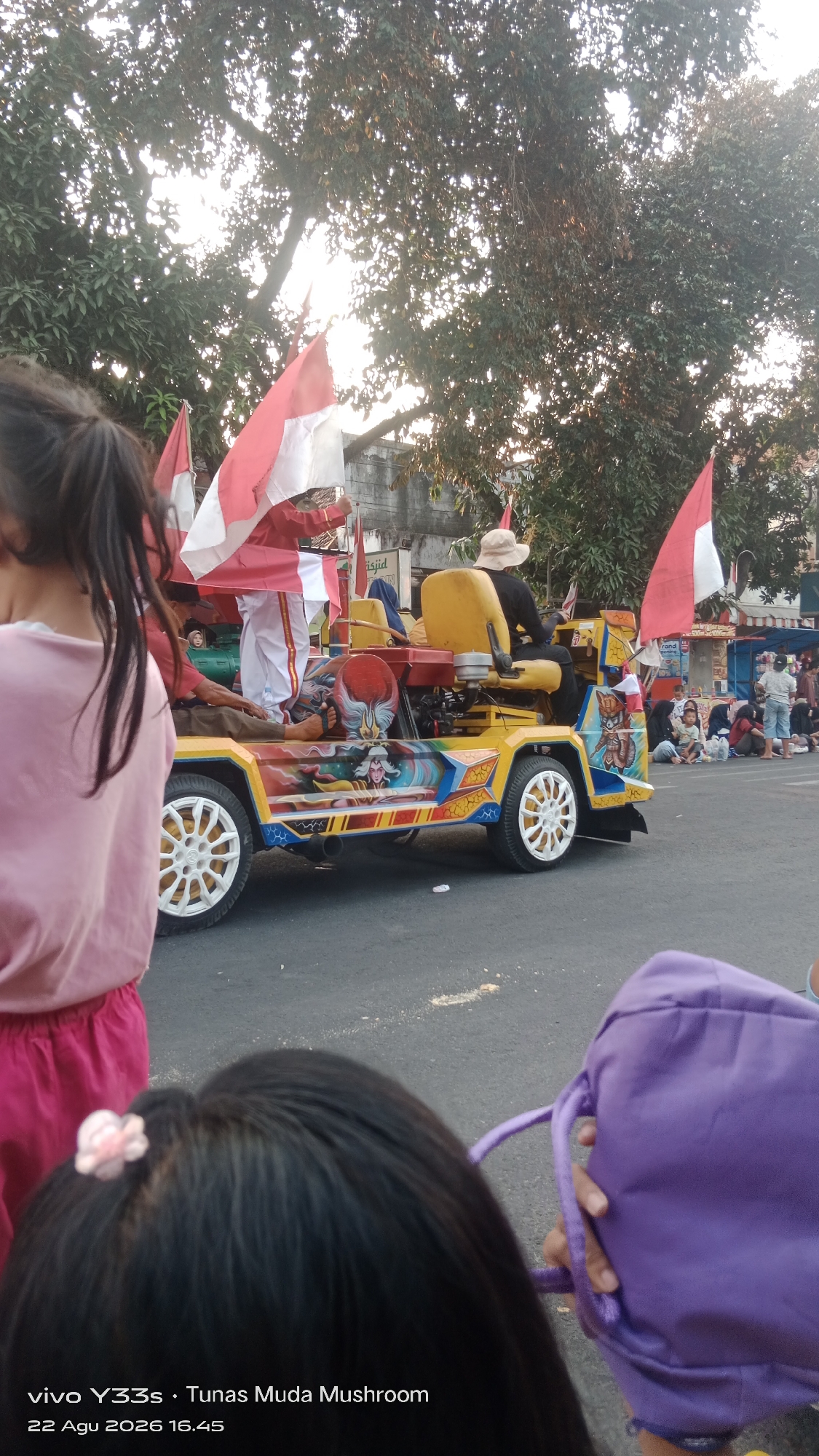 In Genengan, Indonesia, a colorful, modified jeep decorated with elaborate murals drives down a paved street. Indonesian flags are prominently displayed on poles attached to the jeep. Several people are seated on the jeep, including a person wearing a white hat and a dark shirt driving, and another person dressed in red and white standing and holding a flag. In the foreground, two young girls are visible. One girl, wearing a pink shirt and pink shorts, is facing away from the camera. The other girl, also facing away, has black hair adorned with a small pink flower and is holding a purple bag. The background shows trees, buildings, and other people observing the scene, suggesting a parade or public event. The lighting indicates it is either early morning or late afternoon.