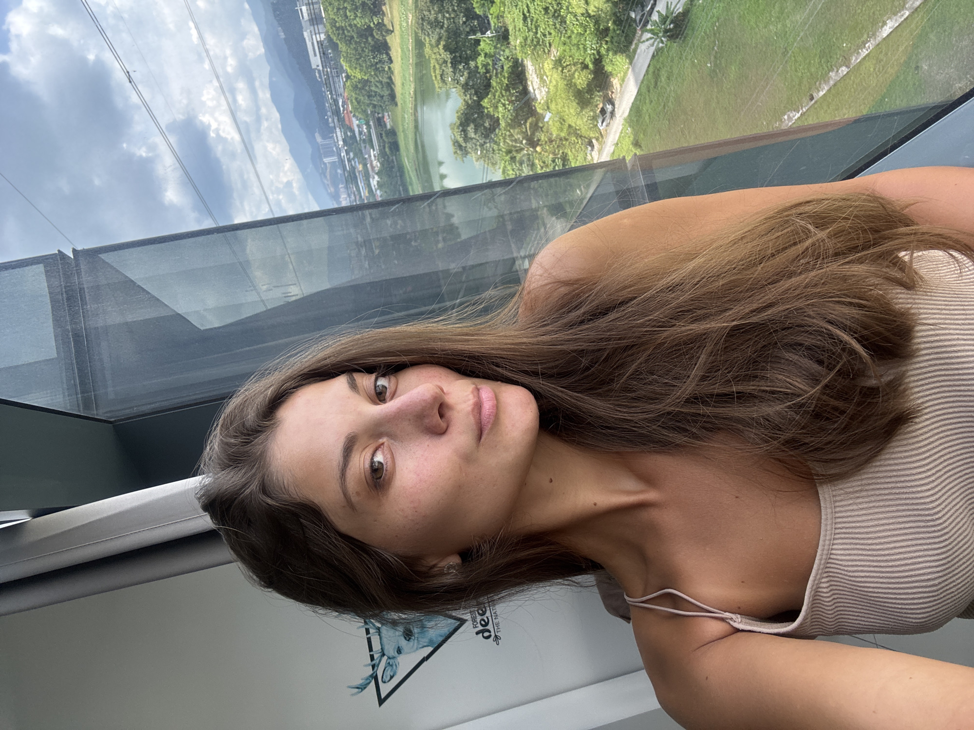 The image captures a young woman taking a selfie indoors, positioned in front of a large glass window or wall. She has long, dark brown hair and is wearing a light beige, ribbed tank top. Her gaze is directed towards the camera with a neutral expression.
The backdrop through the window reveals an expansive outdoor landscape, indicative of a high vantage point overlooking an urban or suburban area with significant natural elements, likely in Kuala Lumpur, Malaysia. A lush green environment, primarily dense trees, is prominent, alongside a body of water, possibly a lake or pond. Further in the distance, buildings of a city or town are visible, nestled against a backdrop of mountains or hills under a partly cloudy sky. Cable lines are noticeable across the sky, suggesting the location could be a cable car or a high observation deck.
The photo was taken during the day, illuminated by natural, diffused light. On an interior surface near the woman's right shoulder, a white sign or sticker features a deer head logo. Visible text on this sign includes "FOREST DEER" and "THE NATURALLY". The image has a slightly tilted perspective, enhancing the dynamic feel of the selfie.