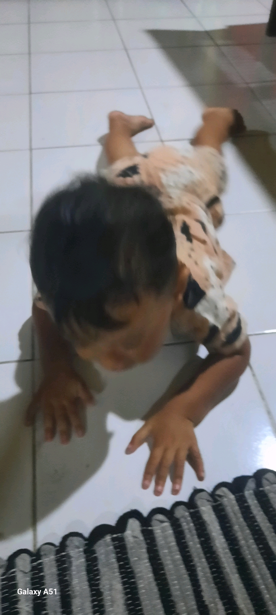 A young child is on all fours on a white tiled floor, likely indoors. The child is wearing a light-colored garment with dark splotches. Their head is bowed, and their hands are placed on the floor in front of them. The lighting suggests an indoor environment with shadows cast on the floor. The lower right corner of the image shows a portion of a black and gray striped rug. The "Galaxy A51" watermark indicates the device used to capture the image. The image is taken in Ploso, Indonesia.