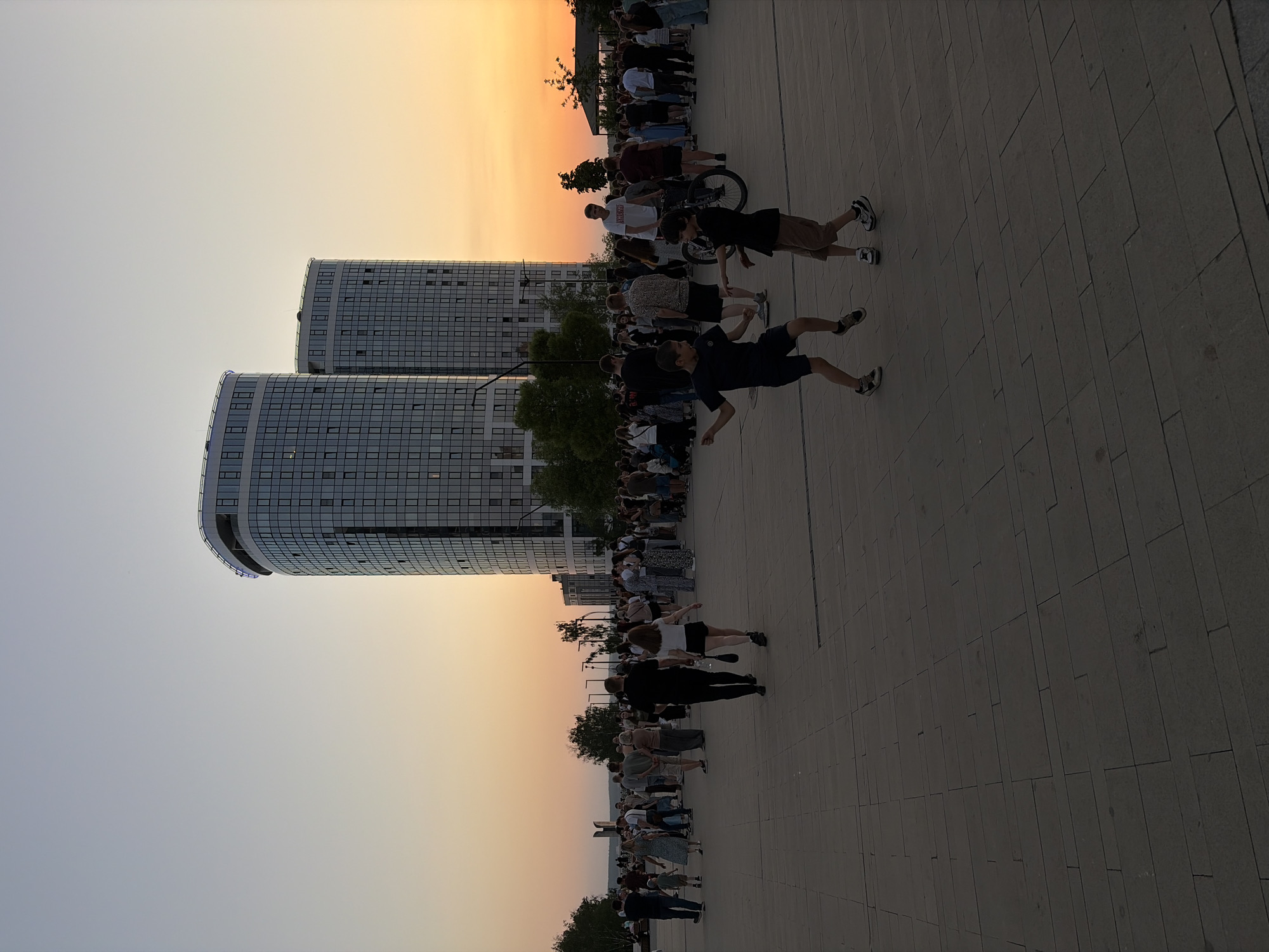 The image depicts a sunset scene in Izhevsk, Russia, featuring two modern, rounded-top high-rise buildings. In the foreground, a crowd of people is gathered on a paved area. Some individuals are standing, walking, or interacting with others. The sky transitions from light orange near the horizon to a pale blue towards the top.