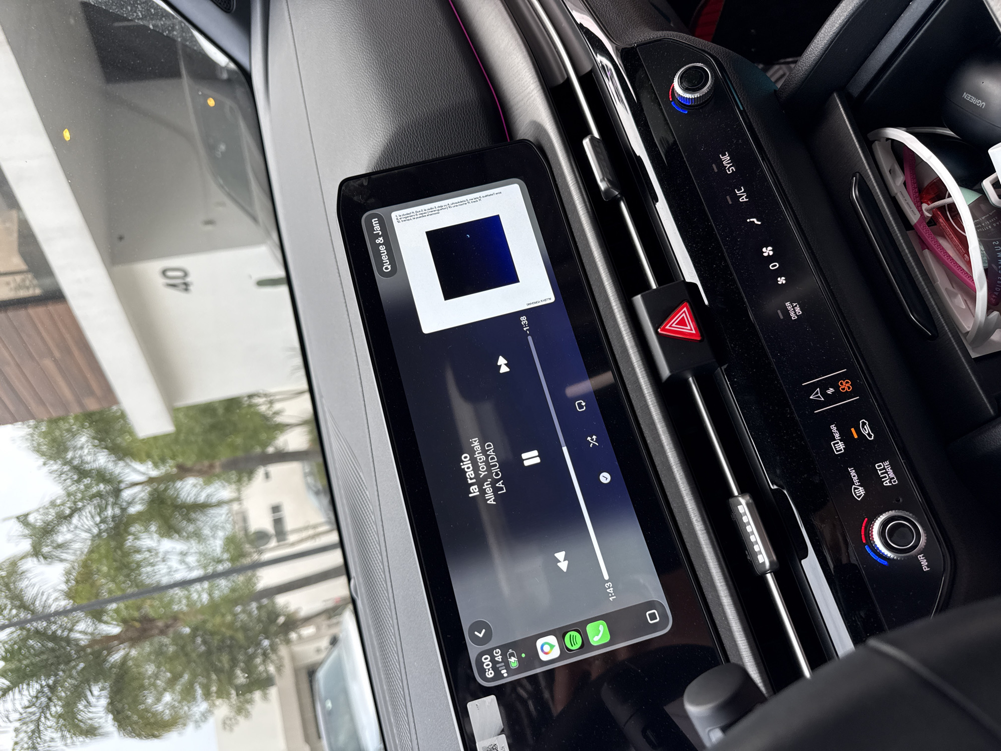 Here is a detailed description of the image:
**Scene:** The image appears to be taken from inside a vehicle, focusing on the dashboard area. The angle is slightly tilted, giving a view of the infotainment system, climate control panel, and some of the exterior environment through the windshield.
**Objects:**
* **Infotainment System:** The central element is a touchscreen displaying a music player interface. It shows a song titled "la radio" by Alleh, Yorghaki from the album "LA CIUDAD" playing on Spotify. There is also an overlay called "Queue & Jam."
* **Climate Control Panel:** To the right of the screen, there's a climate control panel. This includes buttons for A/C sync, temperature control knobs, and controls for front/rear defrost, and driver only setting.
* **Hazard Light Button:** A red triangular hazard light button is visible between the touchscreen and the climate control panel.
* **Dashboard Detailing:** The dashboard shows gray, black and pink accent colors.
* **Other objects:** A UGREEN accessory is visible, along with other personal items.
* **Exterior:** Through the windshield, a building or structure with the number "40" is visible. There are also trees and what seems to be another vehicle.
**Environment:** The time is 6:00.
**Location:** The location in the city of Santa Clara Ocoyucan, Mexico cannot be determined from the image.